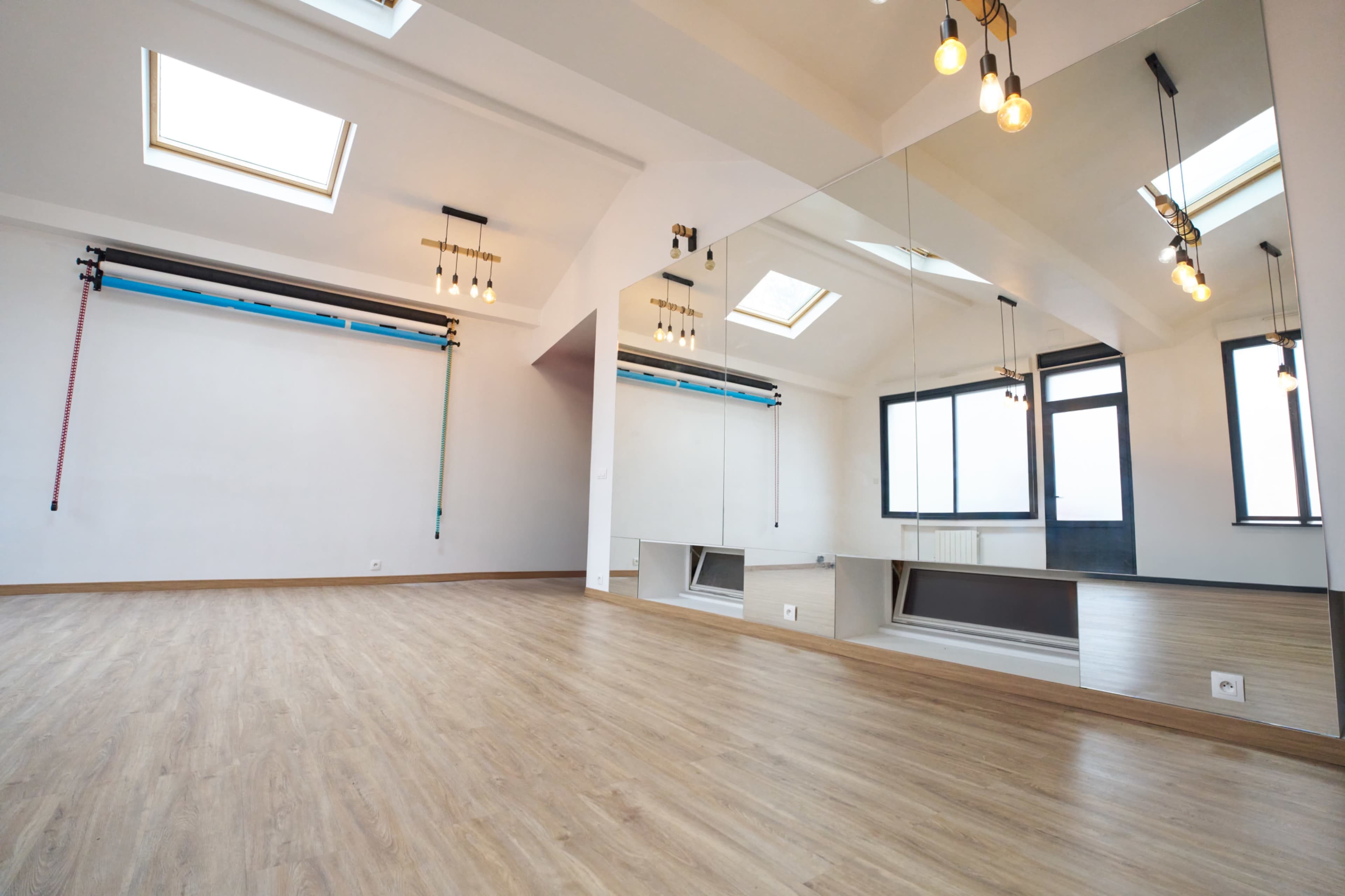 The image shows a spacious, empty room with wooden flooring, a large mirror, and skylights for natural light.
