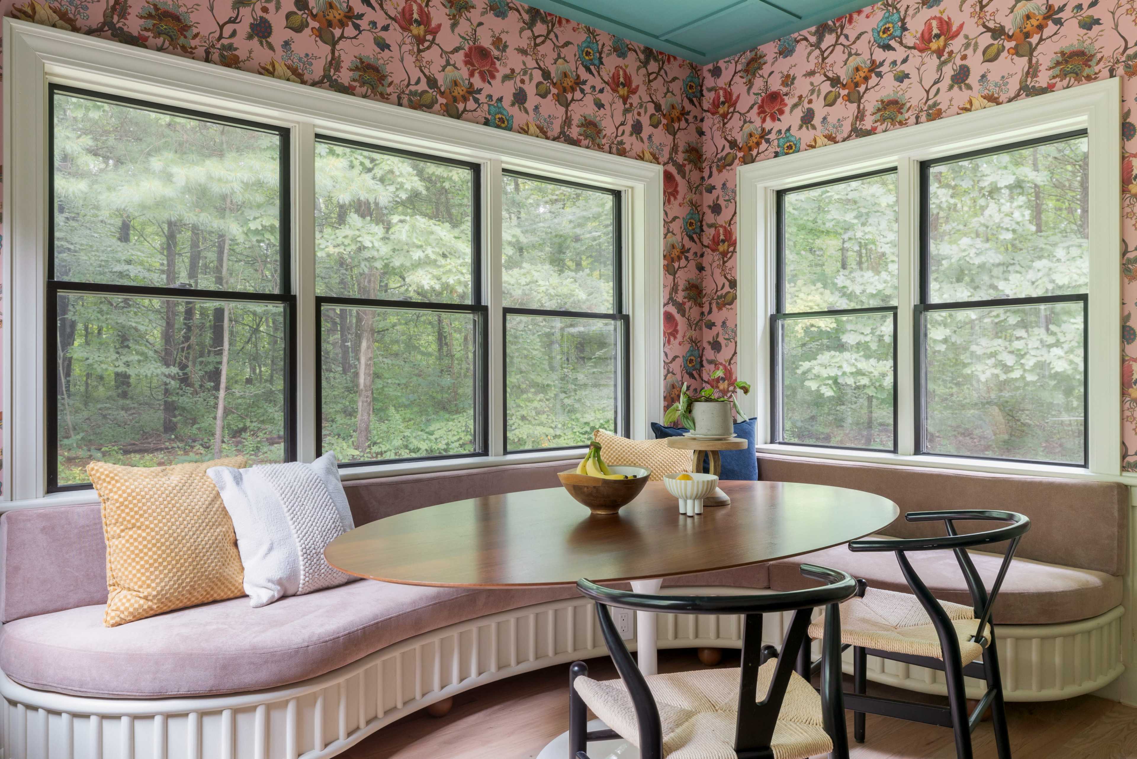 A small dining nook features a curved bench with floral-patterned wallpaper and large windows overlooking a forested area.