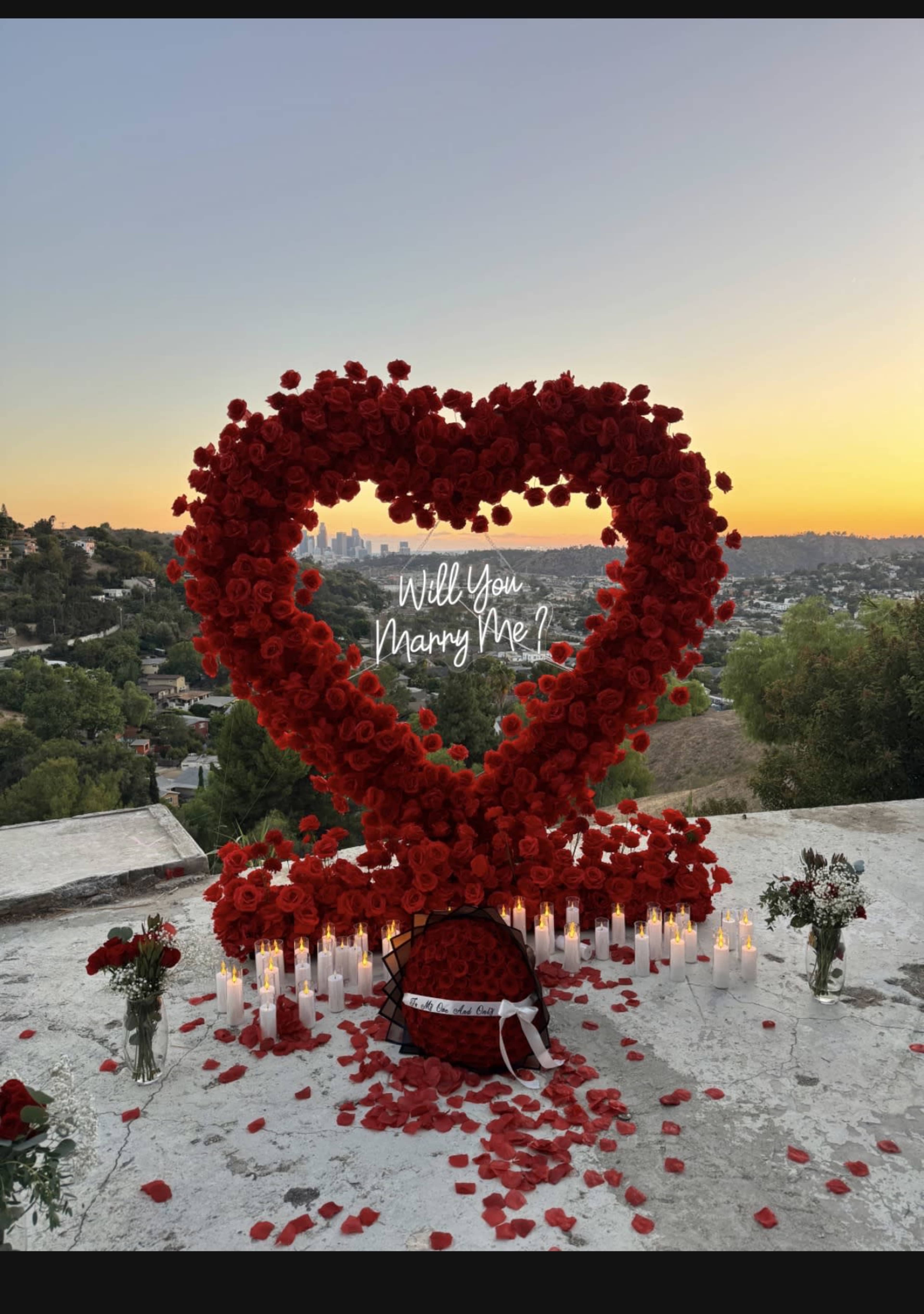 The image shows a large heart-shaped arrangement of red roses surrounded by candles and scattered rose petals, set against a sunset view of a cityscape.