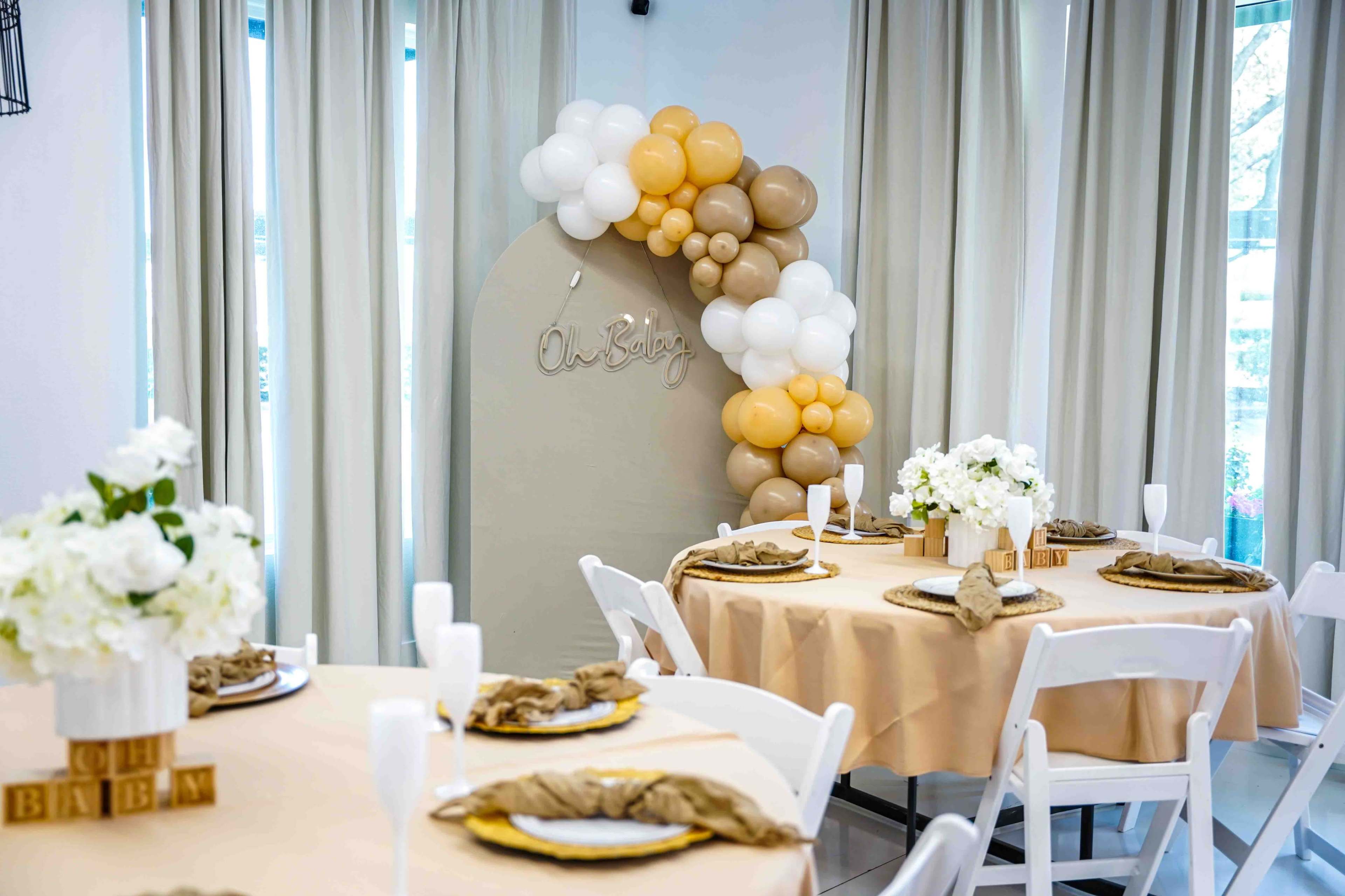 The image shows a decorated event space featuring round tables with beige tablecloths, floral centerpieces, and a balloon arch in neutral tones.