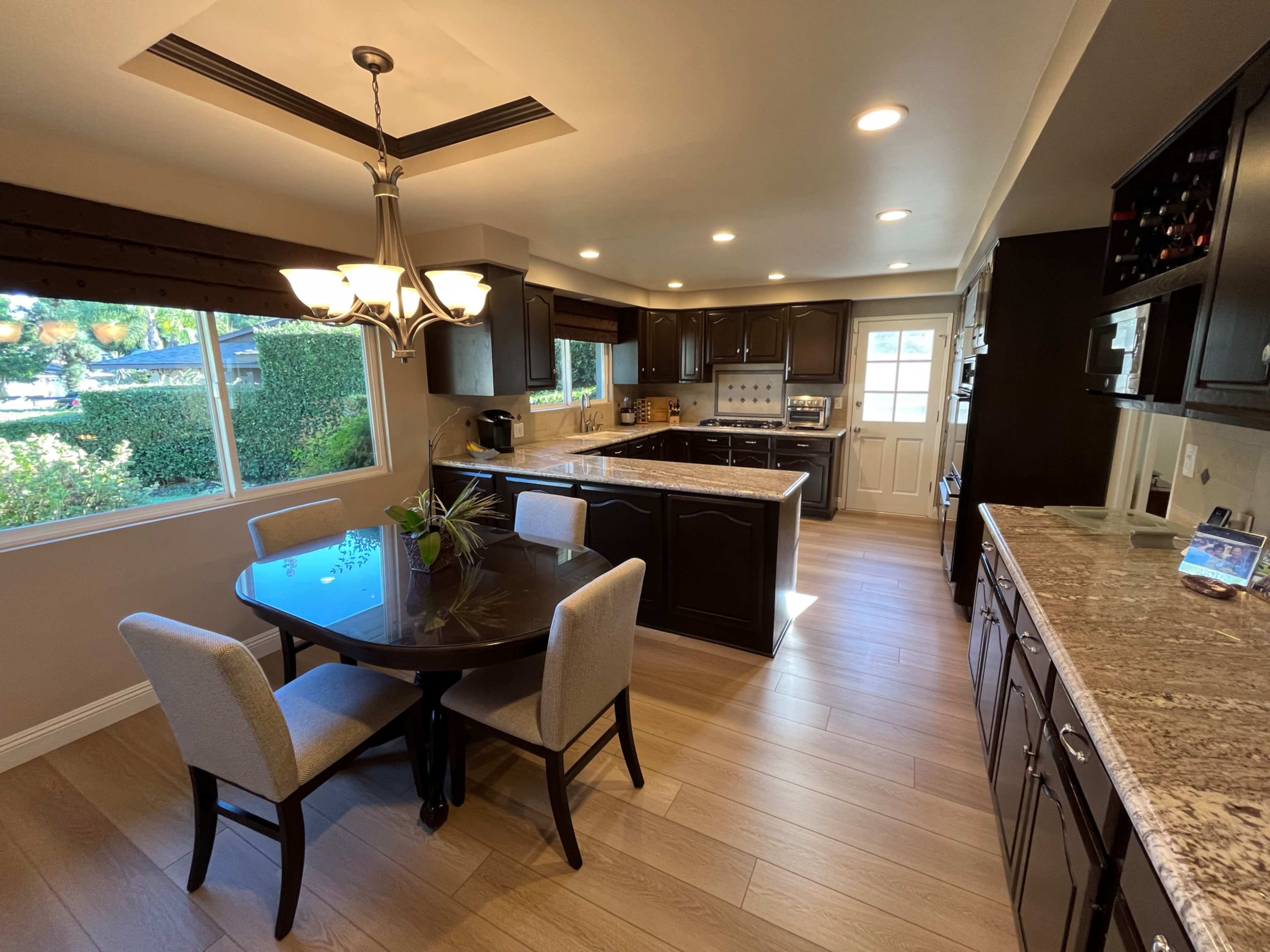 The image shows a modern kitchen with dark wood cabinetry, a granite countertop, a circular dining table with four chairs, and large windows letting in natural light.