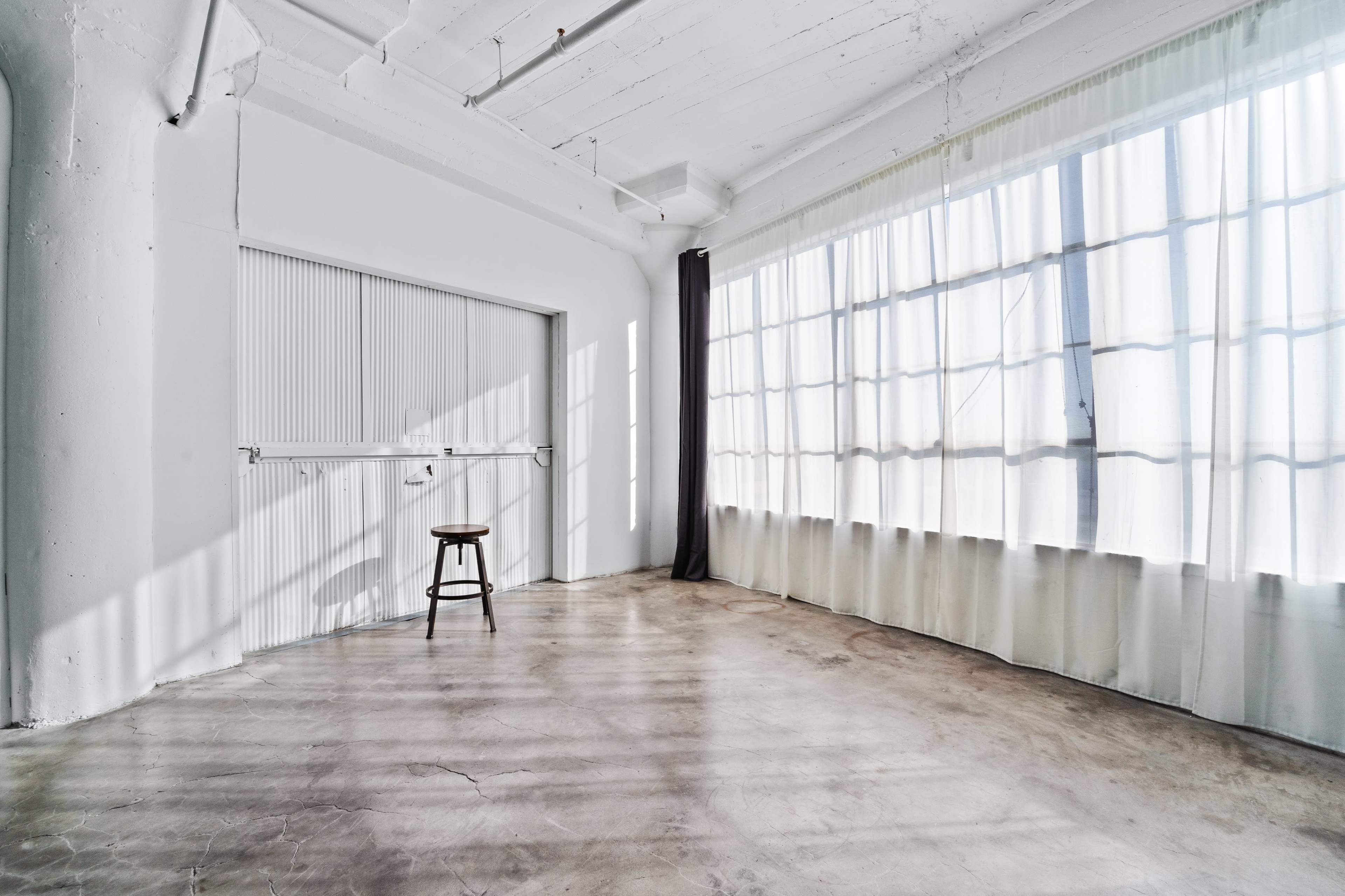 A bright, empty room with large windows, white walls, and a single wooden stool in the center.