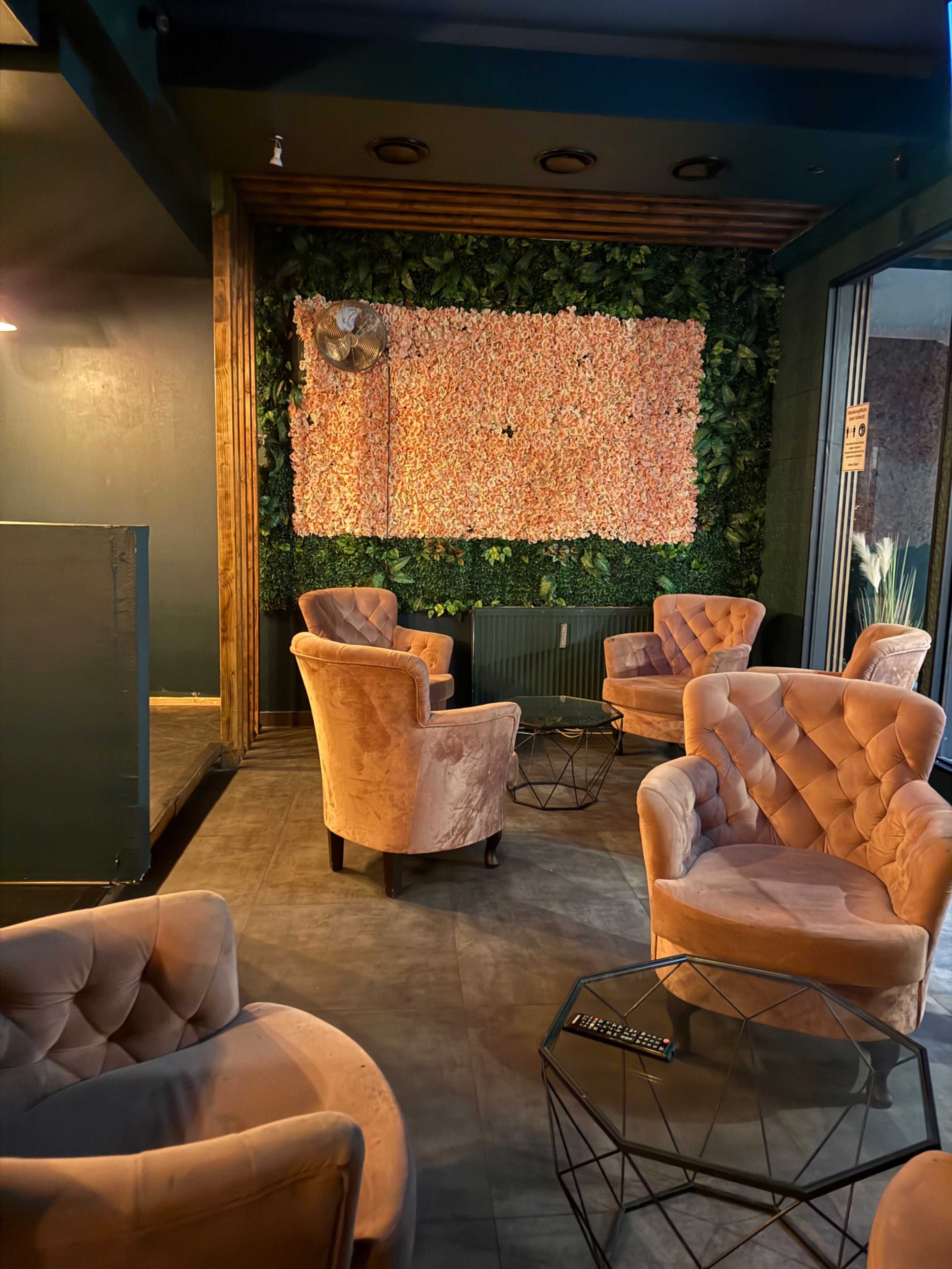 The image shows a cozy seating area featuring several velvet armchairs arranged around small metal tables, set against a backdrop of a floral wall and greenery.