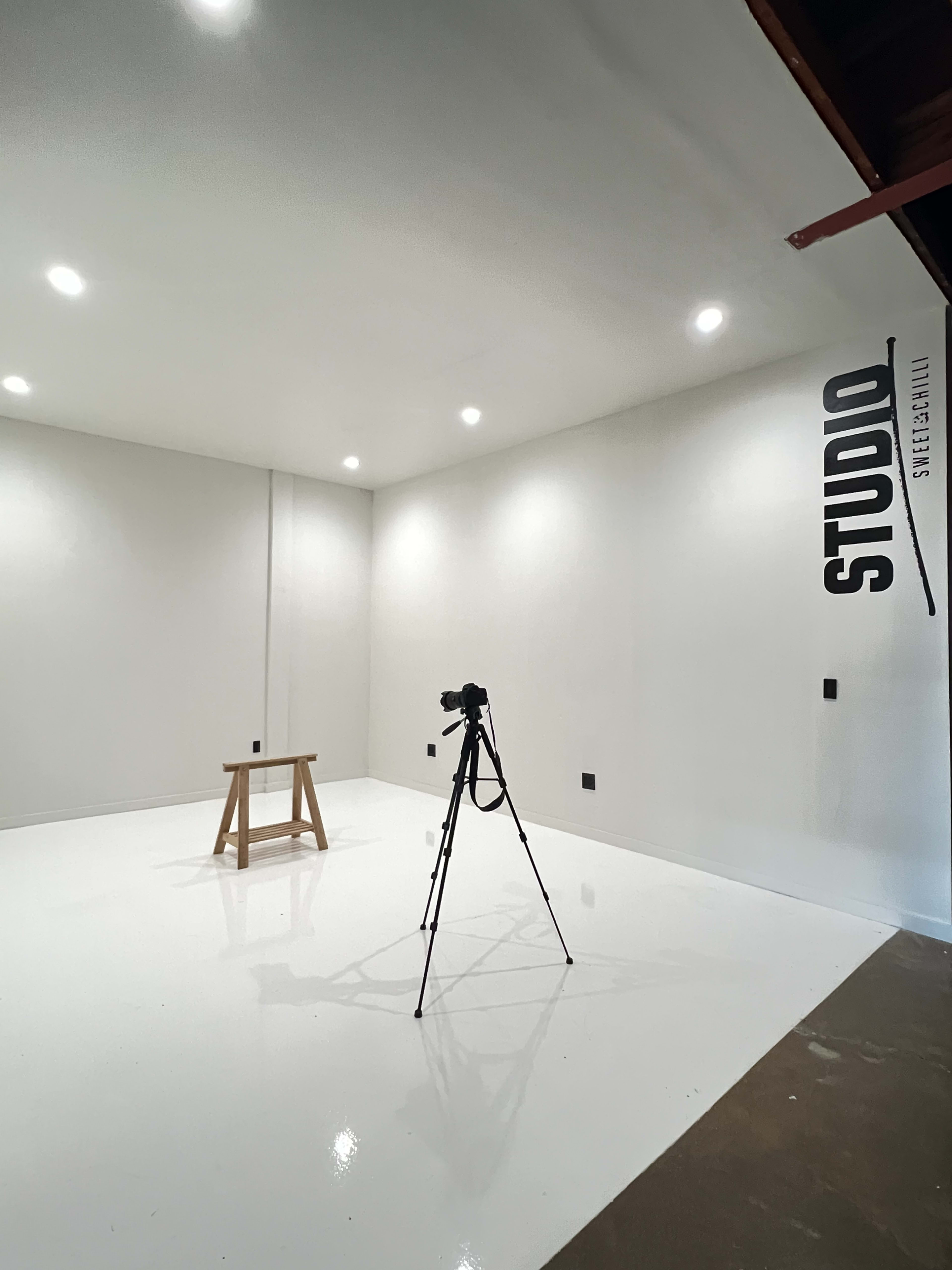 The image shows a minimalistic studio space with a wooden stool and a camera tripod positioned on a white floor under bright lights.