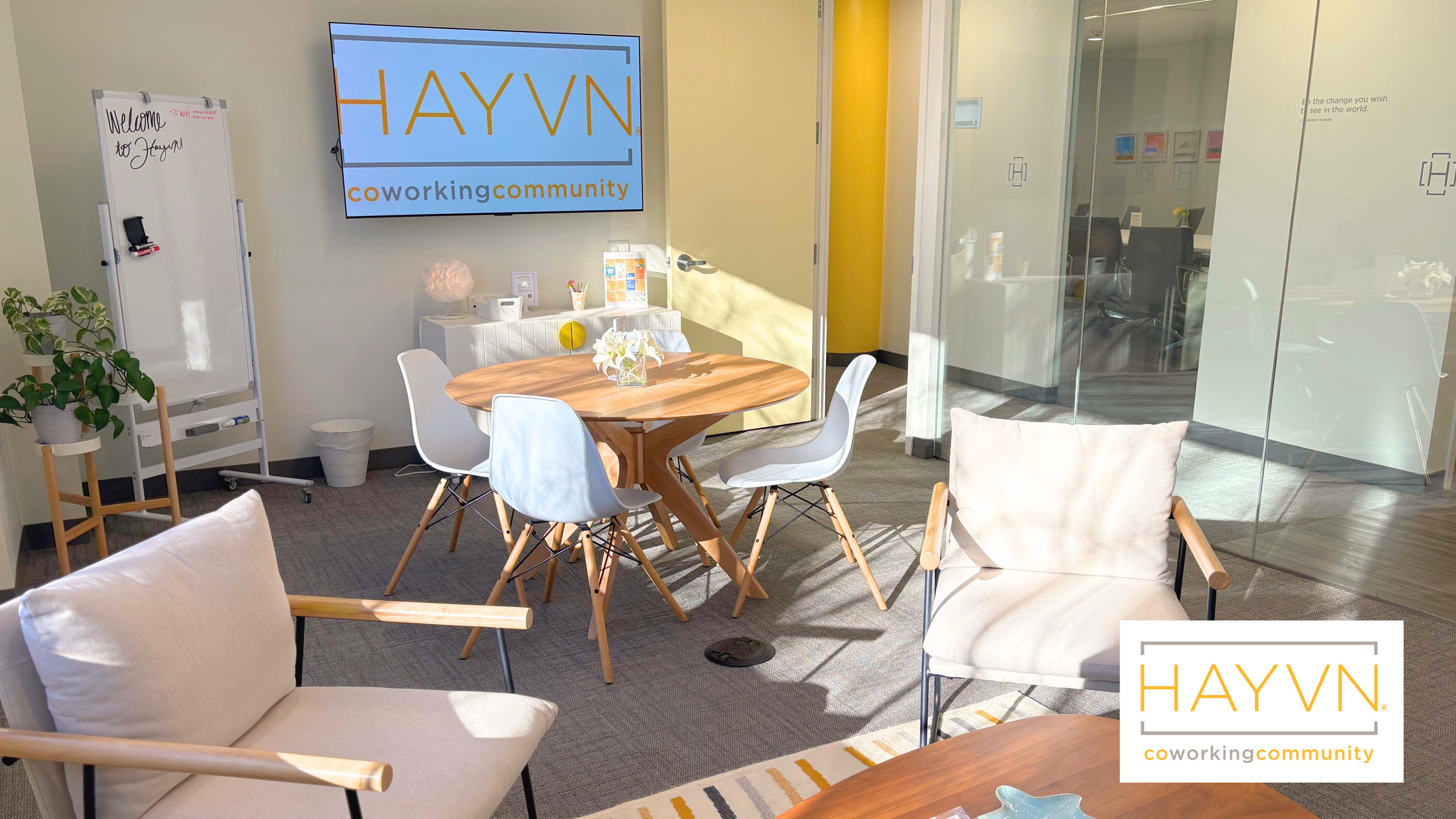 The image shows a bright, modern coworking space featuring a round wooden table, white chairs, and a large screen displaying the HAYVN logo on the wall.
