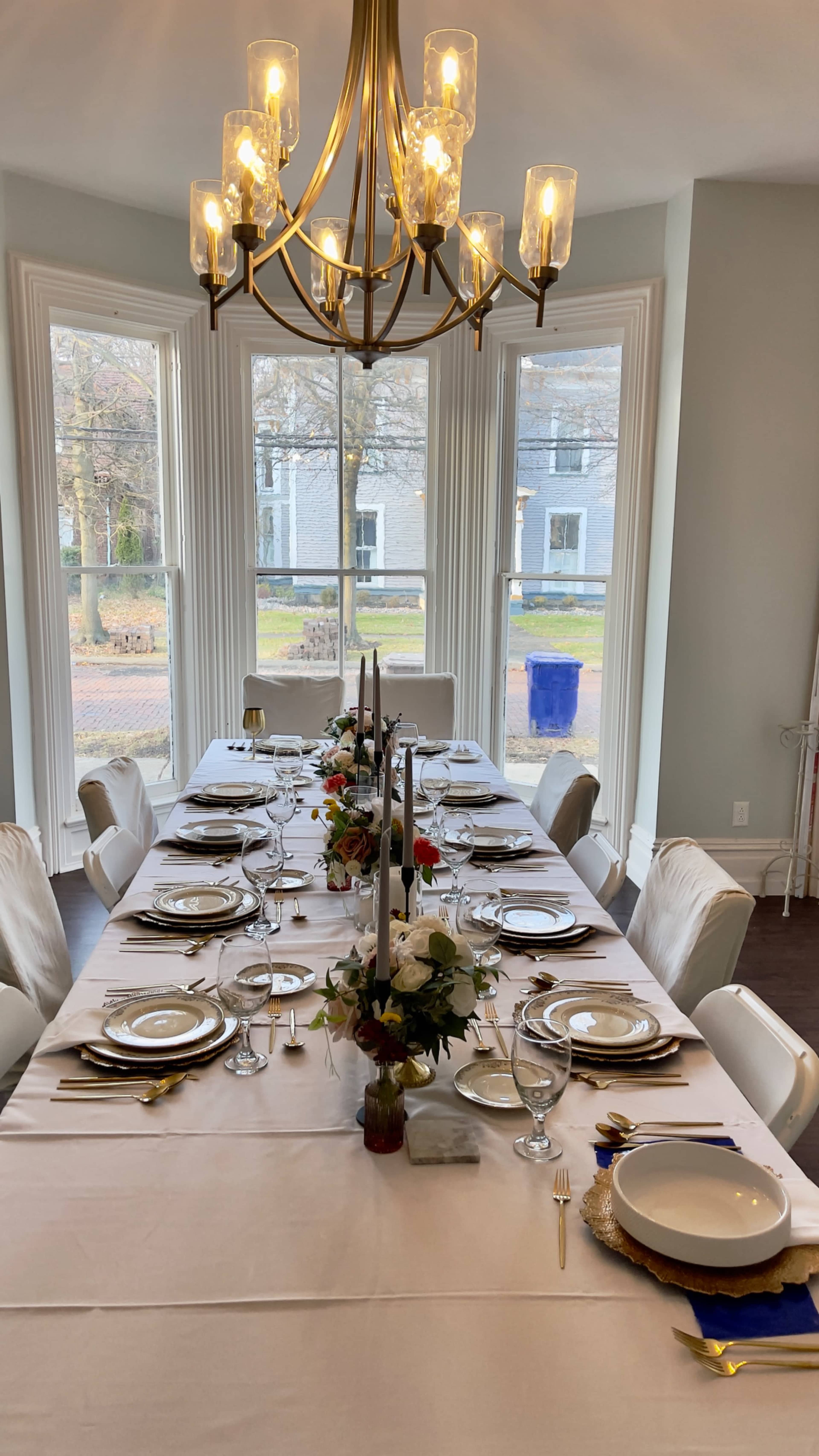 A long dining table is elegantly set with plates, glasses, and a floral centerpiece, positioned under a chandelier in a bright room with large windows.
