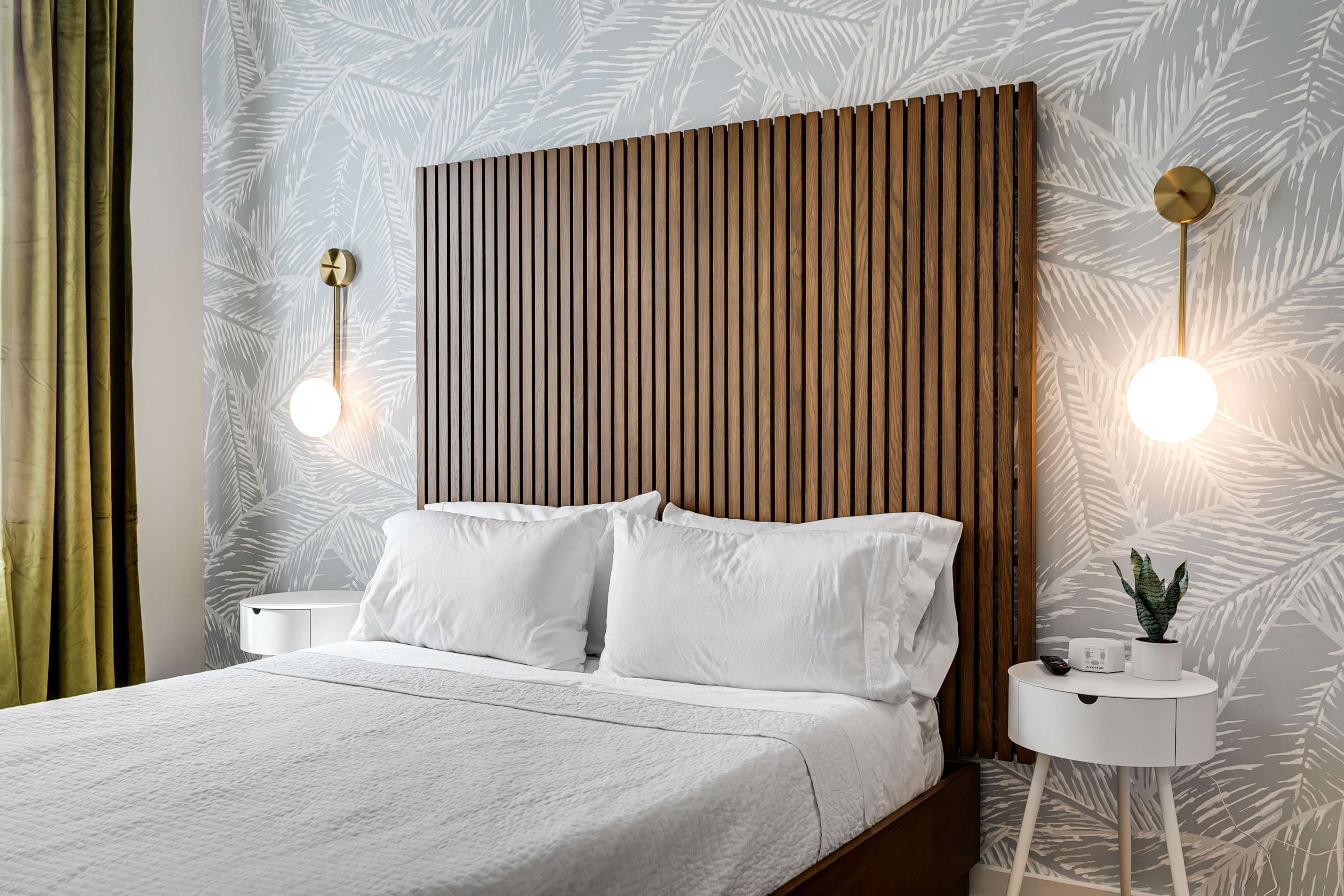 A neatly made bed with white linens is positioned against a wooden slatted headboard, flanked by round wall sconces, in a room featuring patterned wallpaper.