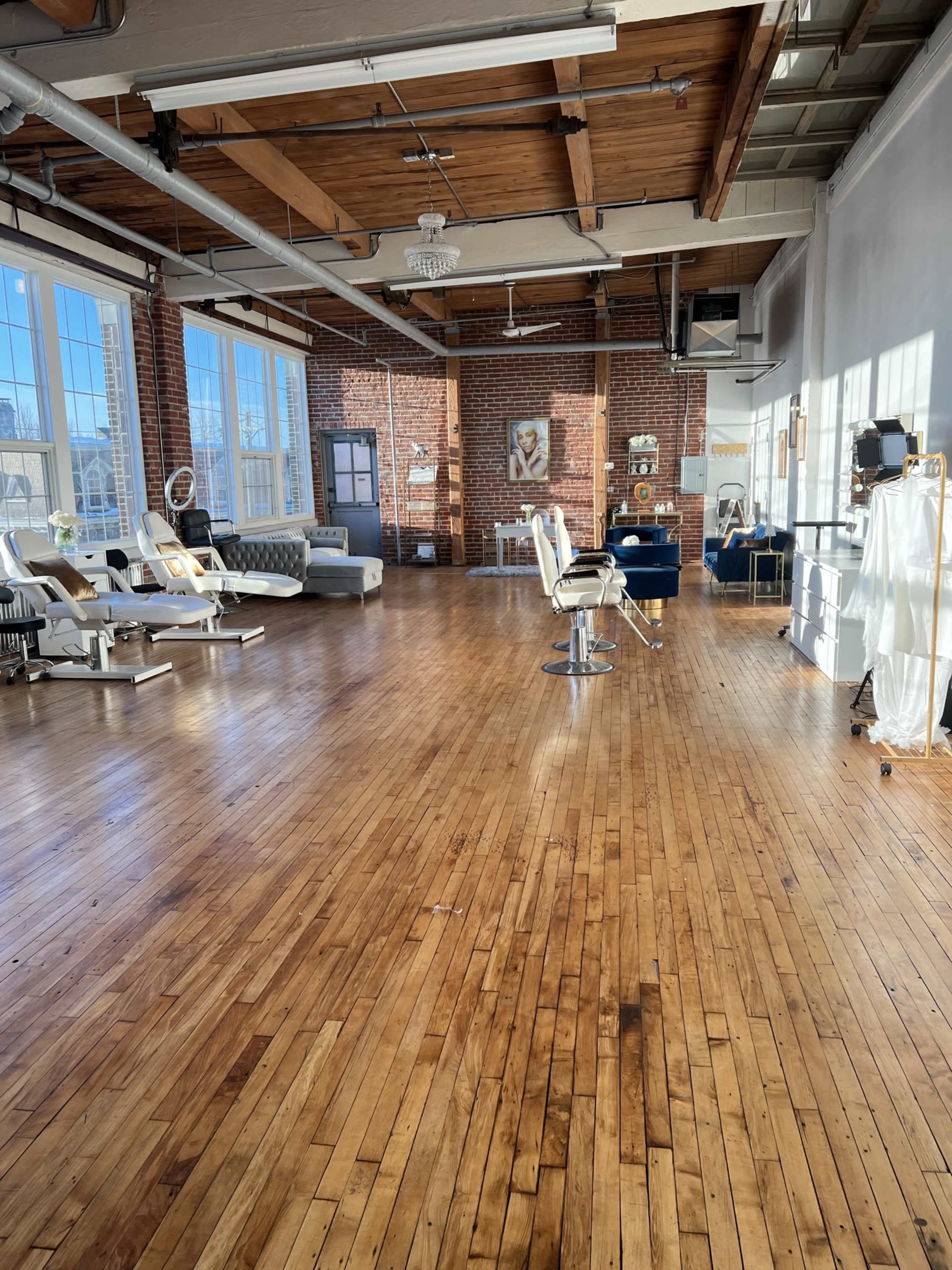 The image shows a spacious, well-lit interior featuring hardwood floors and various pieces of furniture, including salon chairs, a treatment bed, and decorative elements against brick and white walls.