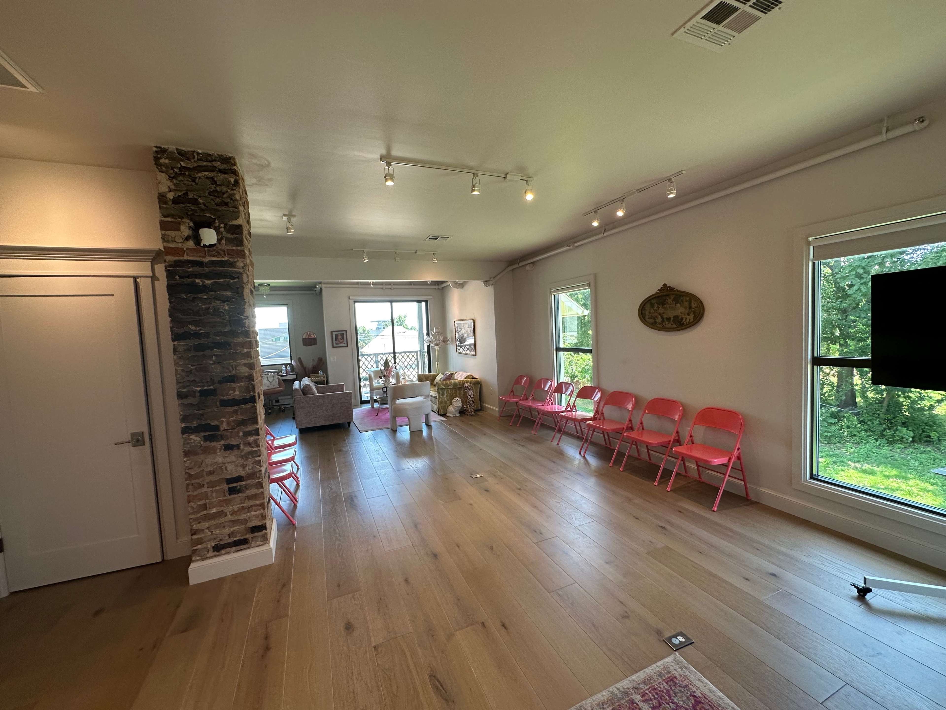 A spacious room with a wooden floor, a seating area with a sofa, and rows of pink chairs arranged along one side, with large windows providing natural light.