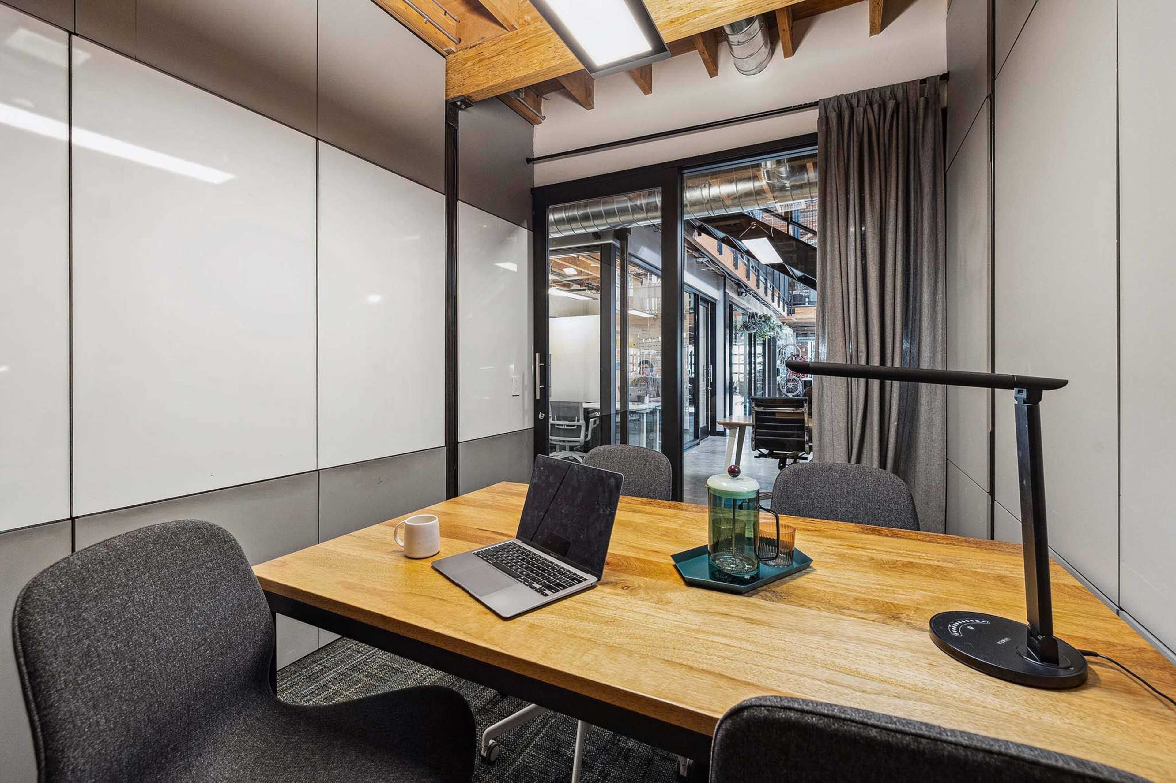 A modern conference room features a wooden table with a laptop, mug, and a decorative tray, surrounded by four chairs.