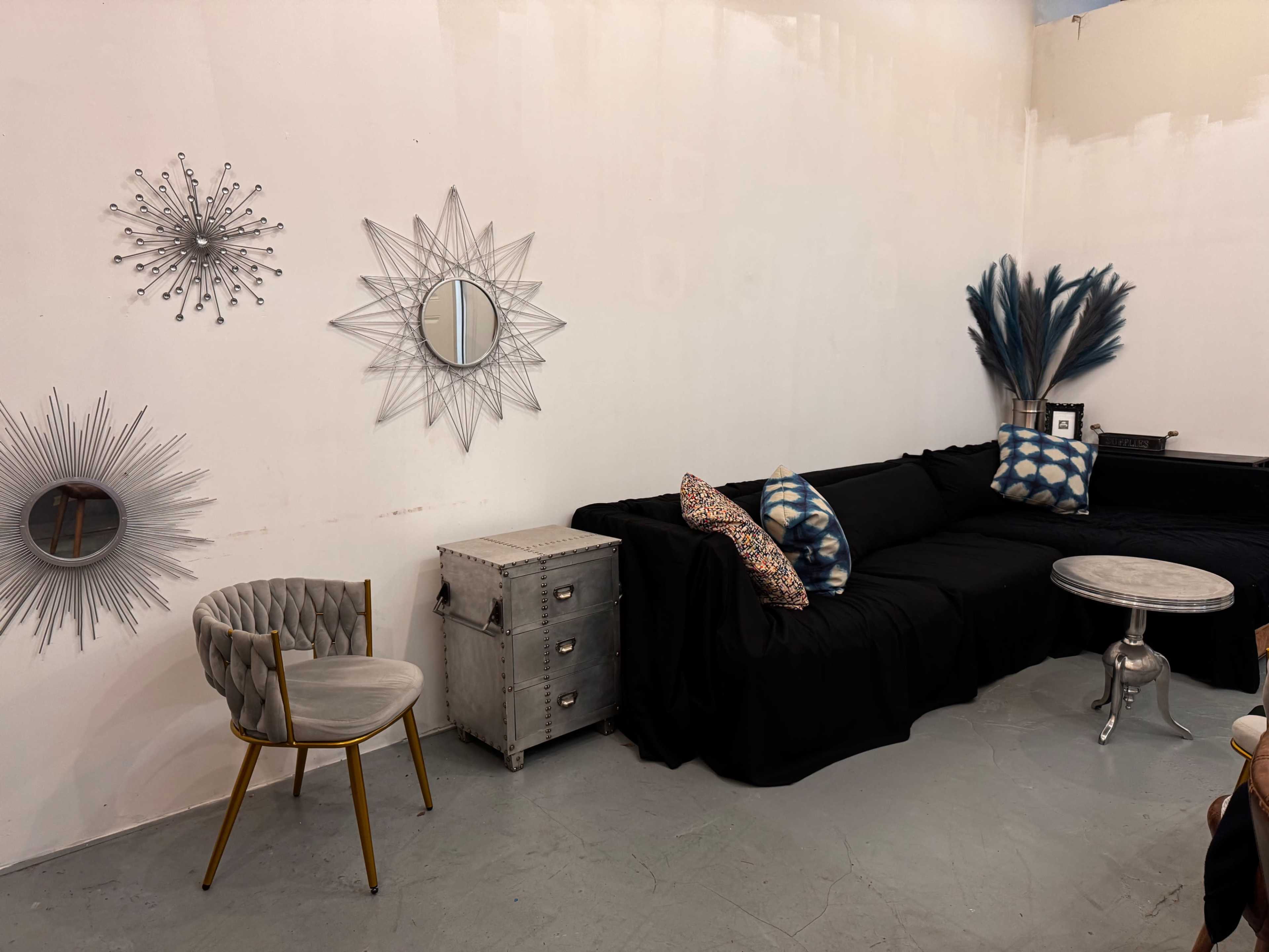 The image shows a modern seating area featuring a black sofa, a round silver table, a tufted chair with gold legs, and decorative mirrors and cushions on the walls.