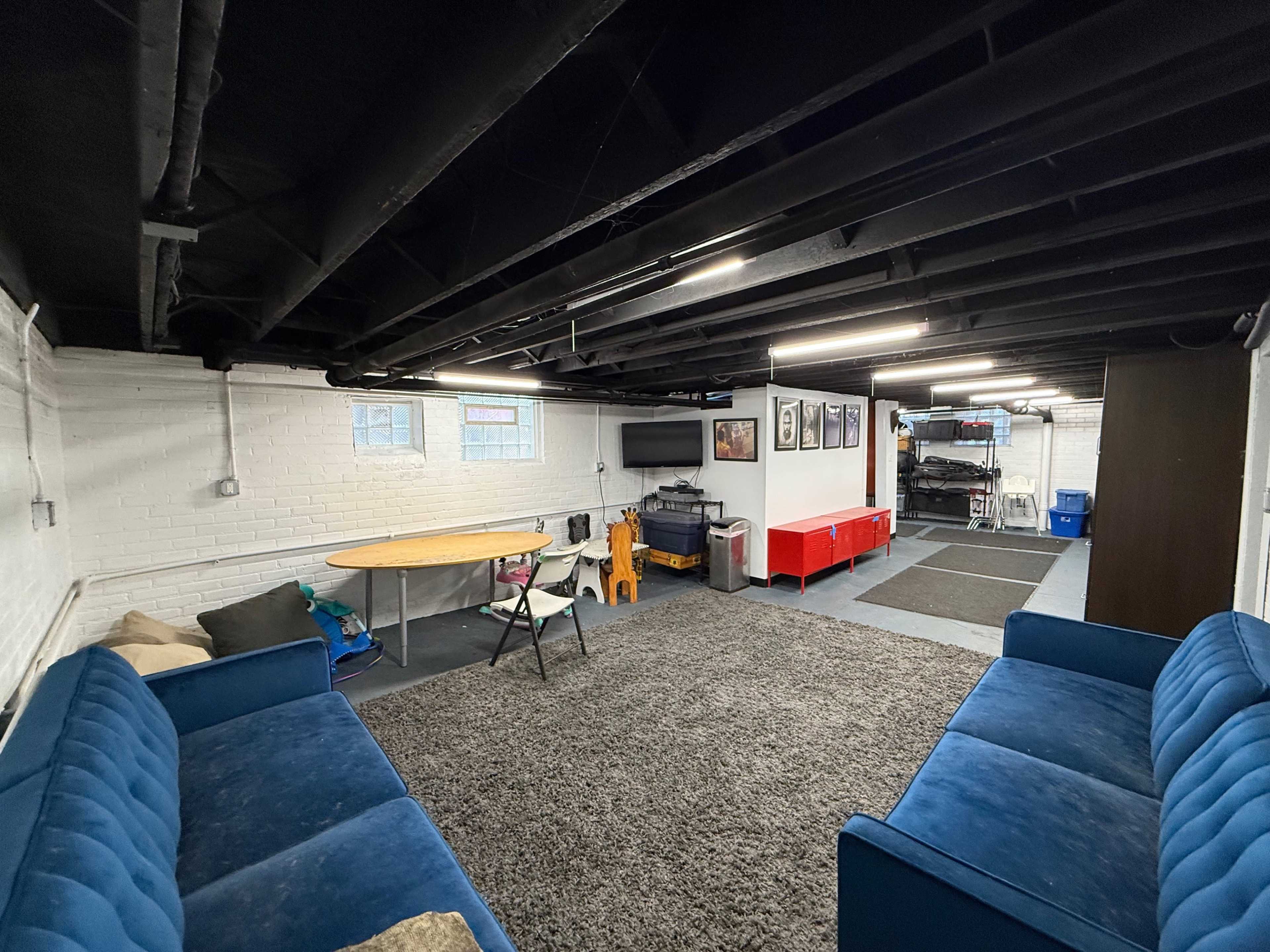The image shows a spacious basement area with two blue sofas, a round table, a television mounted on the wall, and a red storage area in the background.