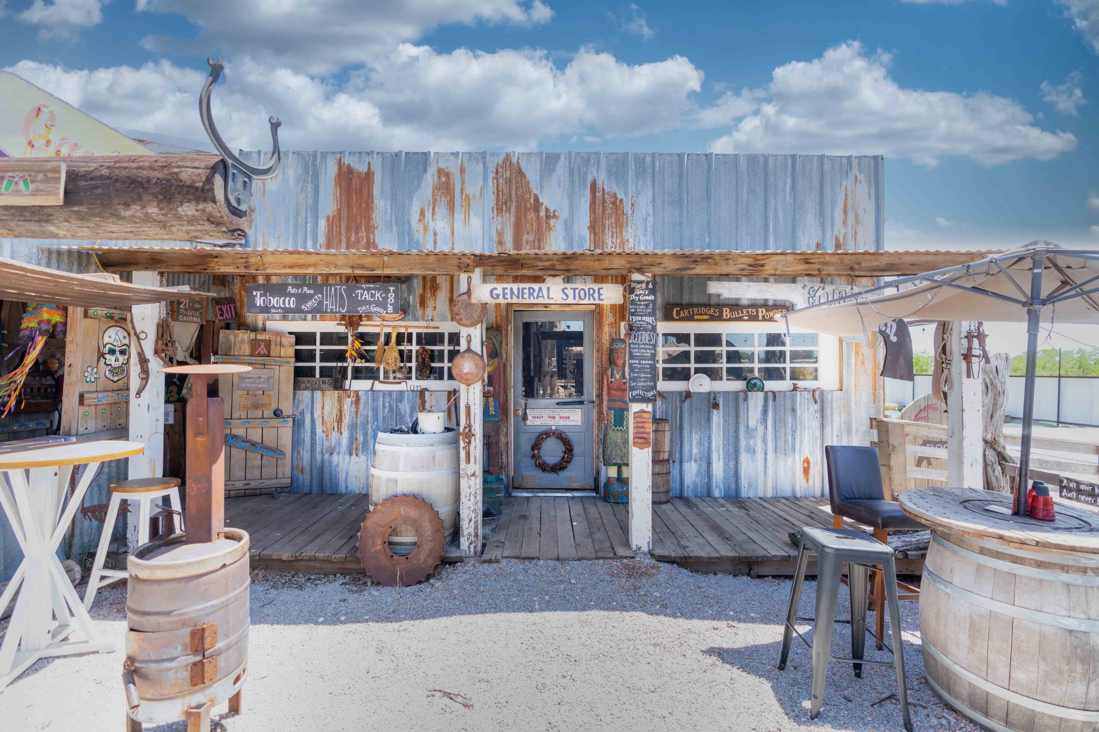 A weathered general store with a wooden deck and rustic decor stands under a blue sky with scattered clouds.