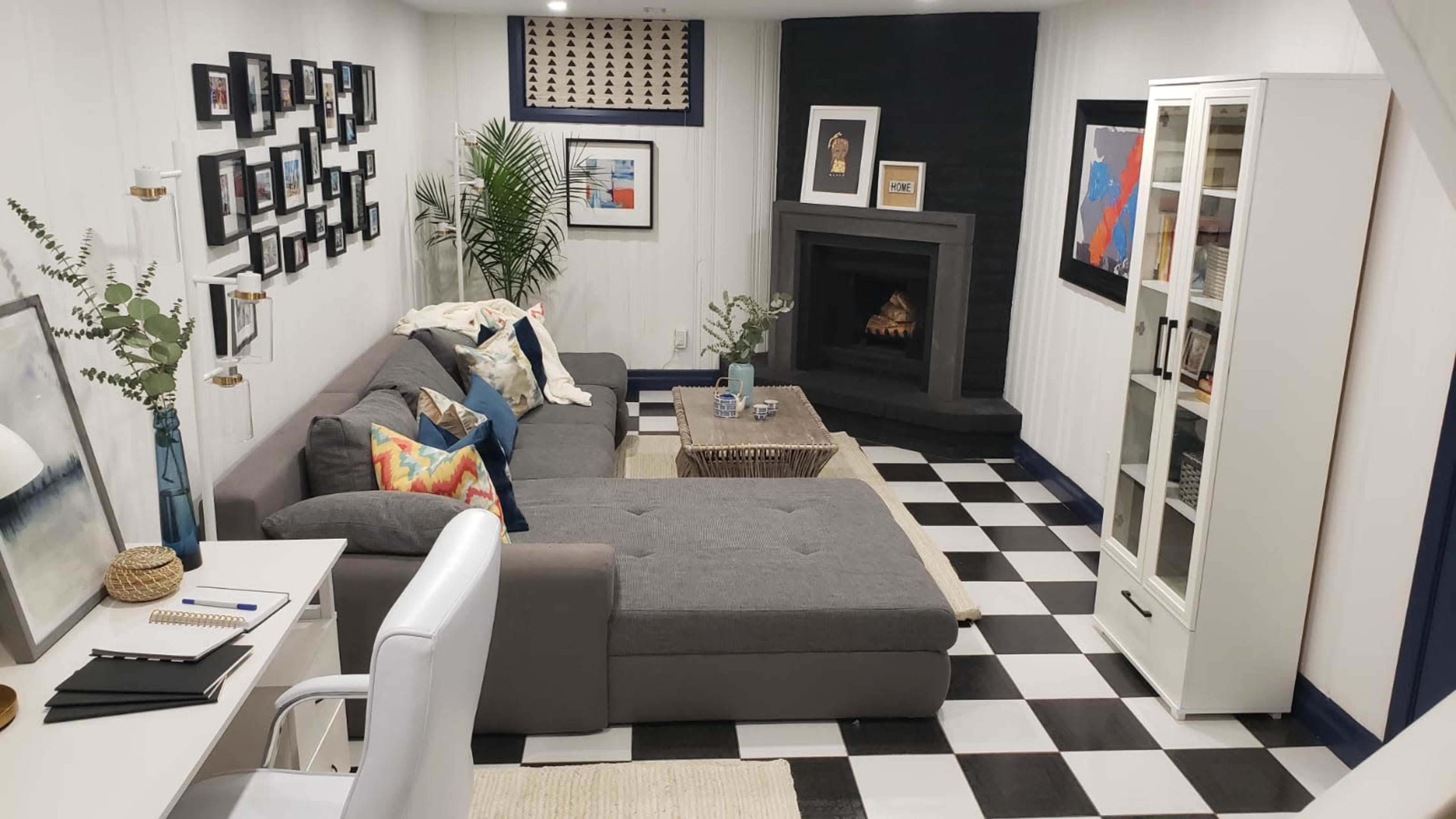 The image shows a modern living room featuring a gray sectional sofa, a fireplace framed by dark tiles, a coffee table, a white bookshelf, and a gallery wall of framed art.