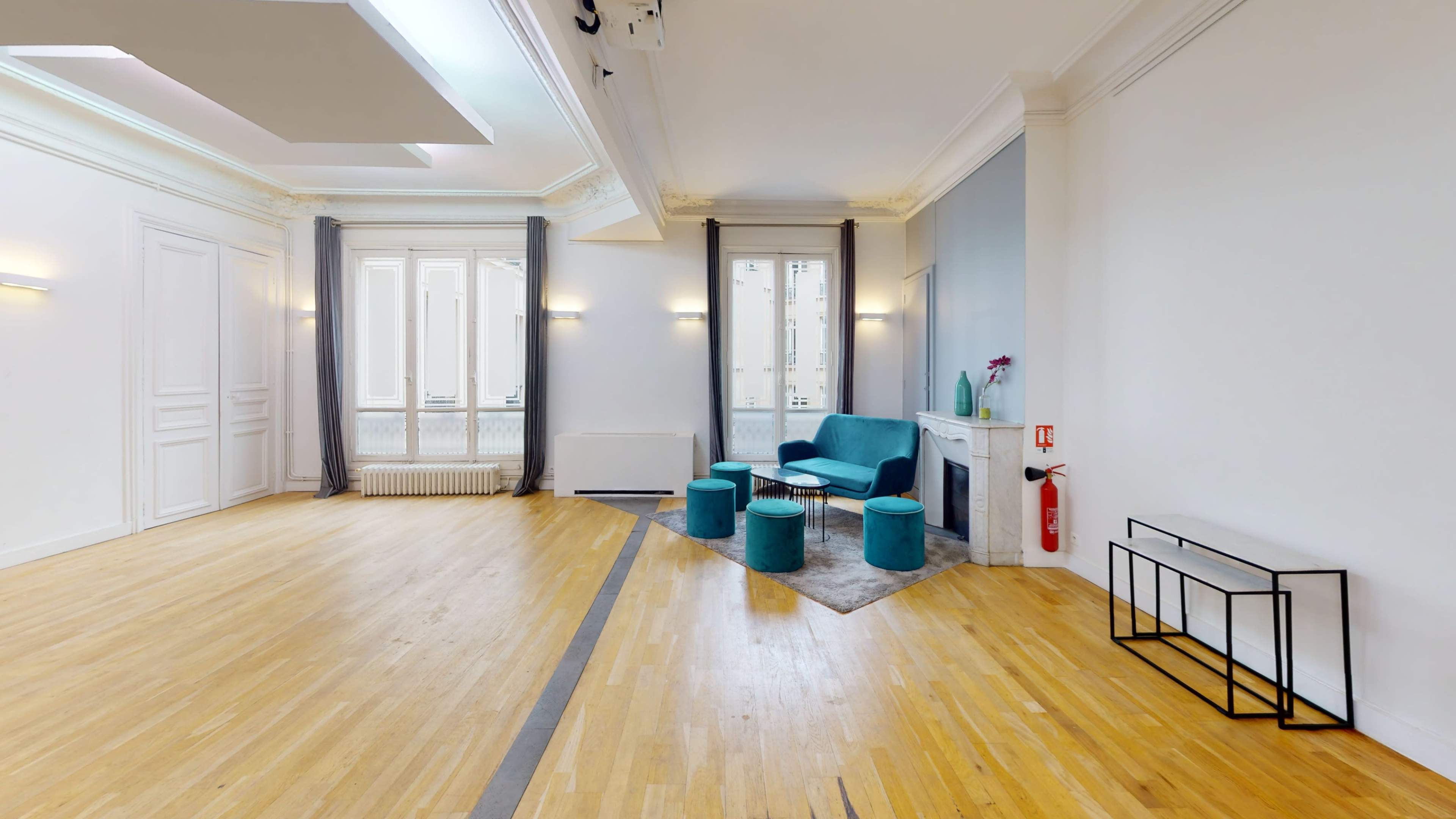 A bright, spacious room with wooden flooring, featuring a seating area with a blue couch and matching poufs, complemented by large windows and elegant light fixtures.