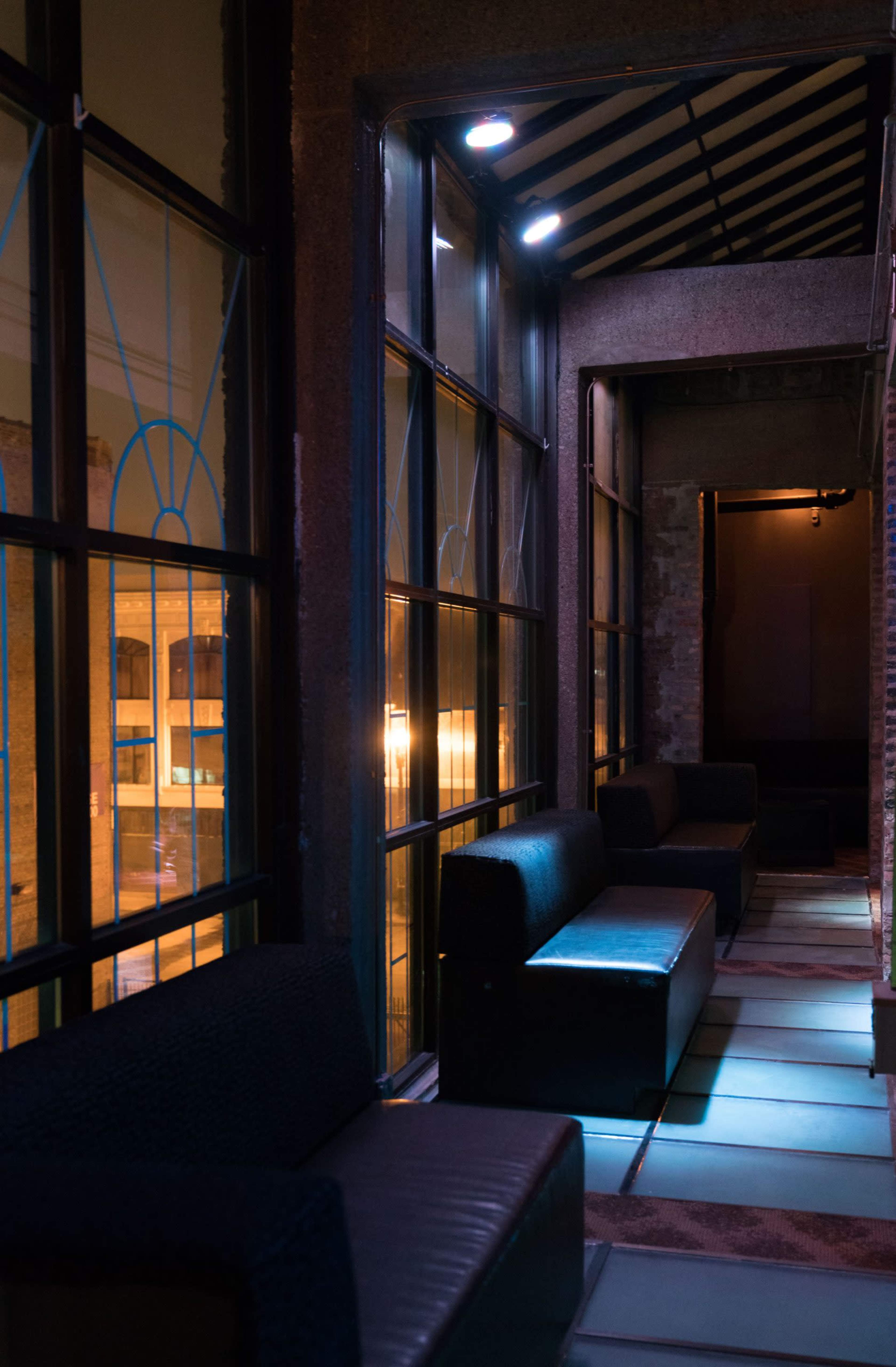 The image features a narrow corridor with sleek black benches and large windows, illuminated by soft blue lighting.