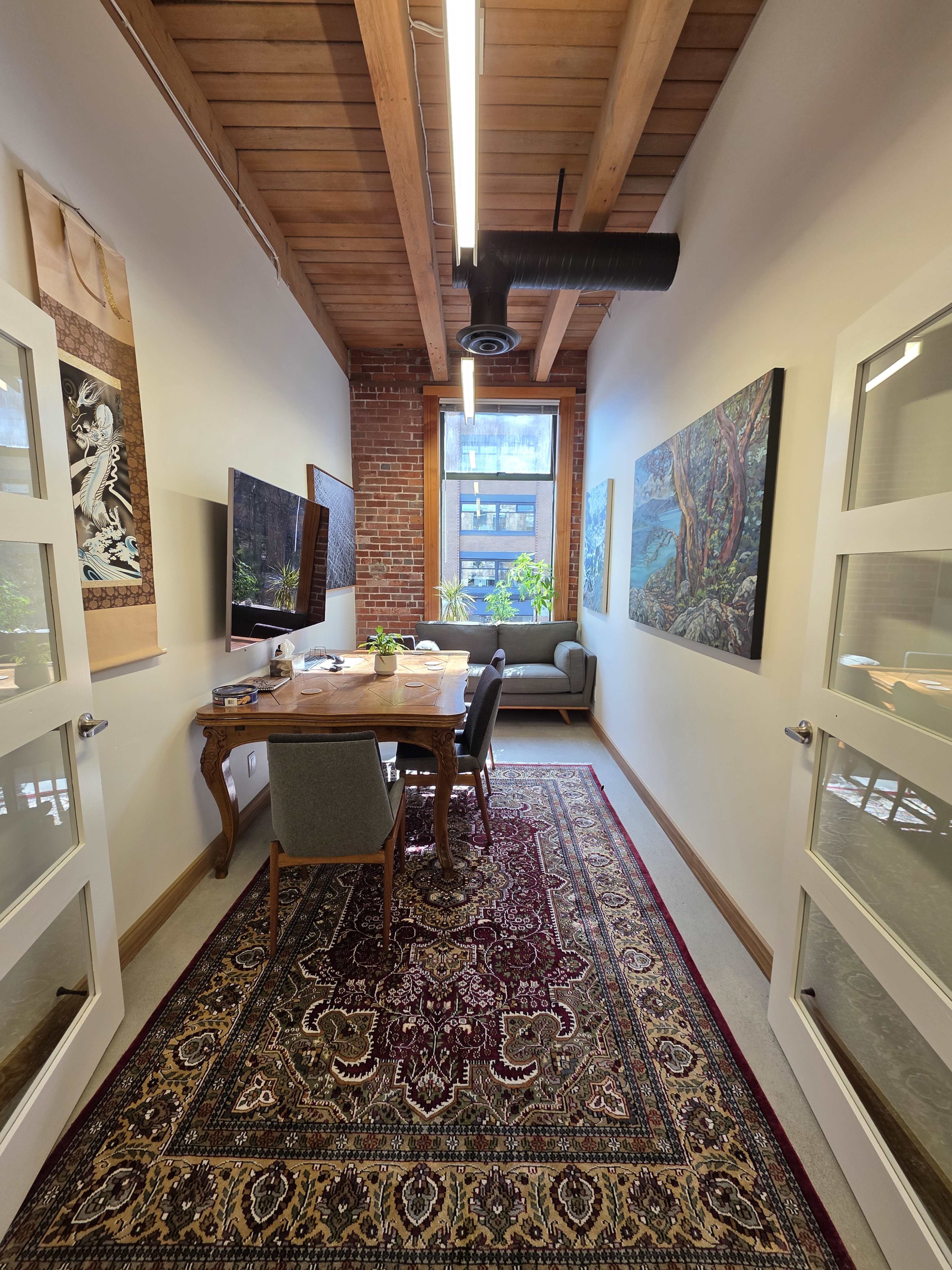 The image shows a narrow office space featuring a wooden table with chairs, a television mounted on the wall, and artwork displayed alongside exposed brick and a decorative rug.