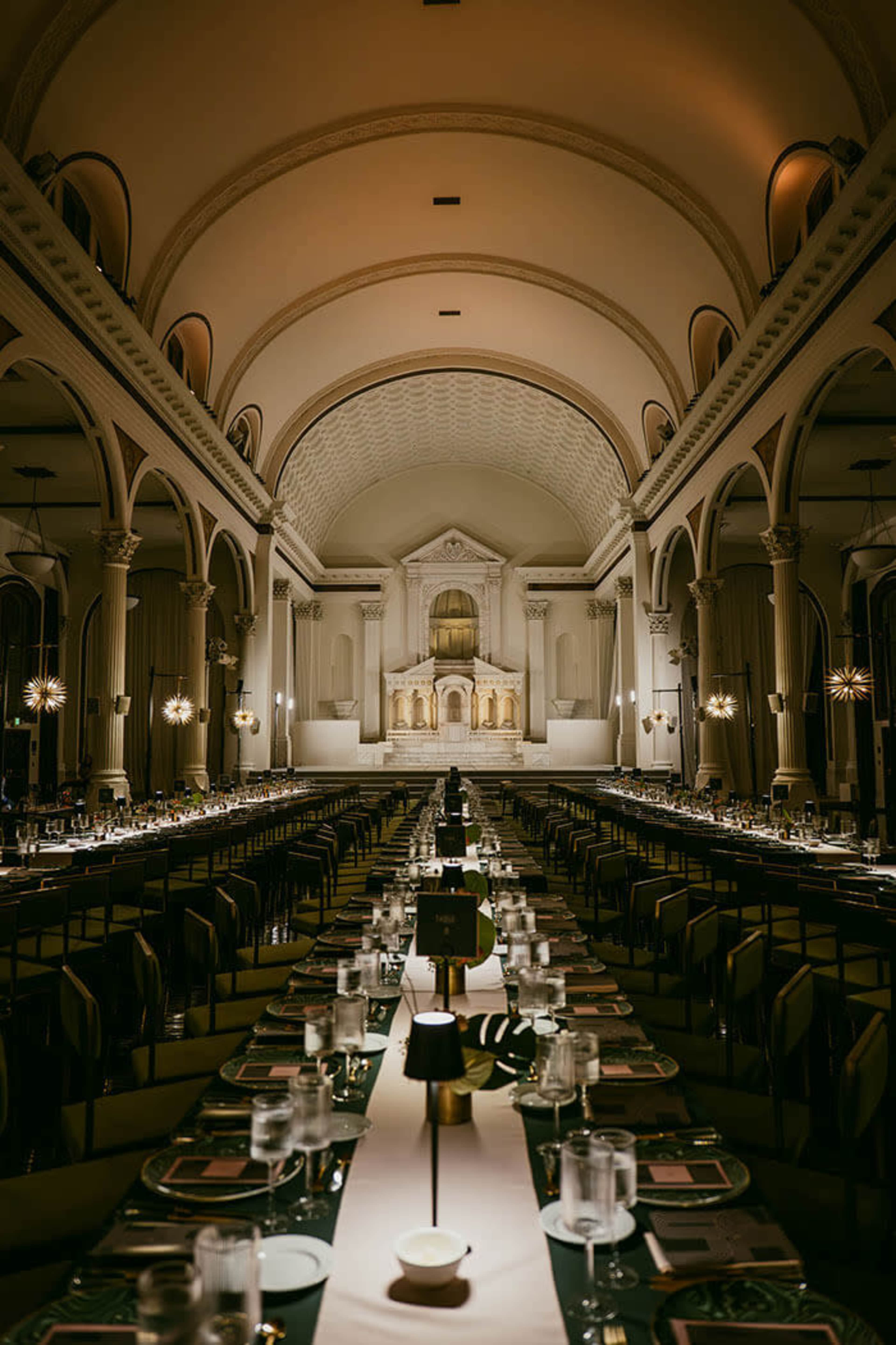 A long dining table is set in a grand hall with high ceilings and ornate architectural details, illuminated by soft lighting.