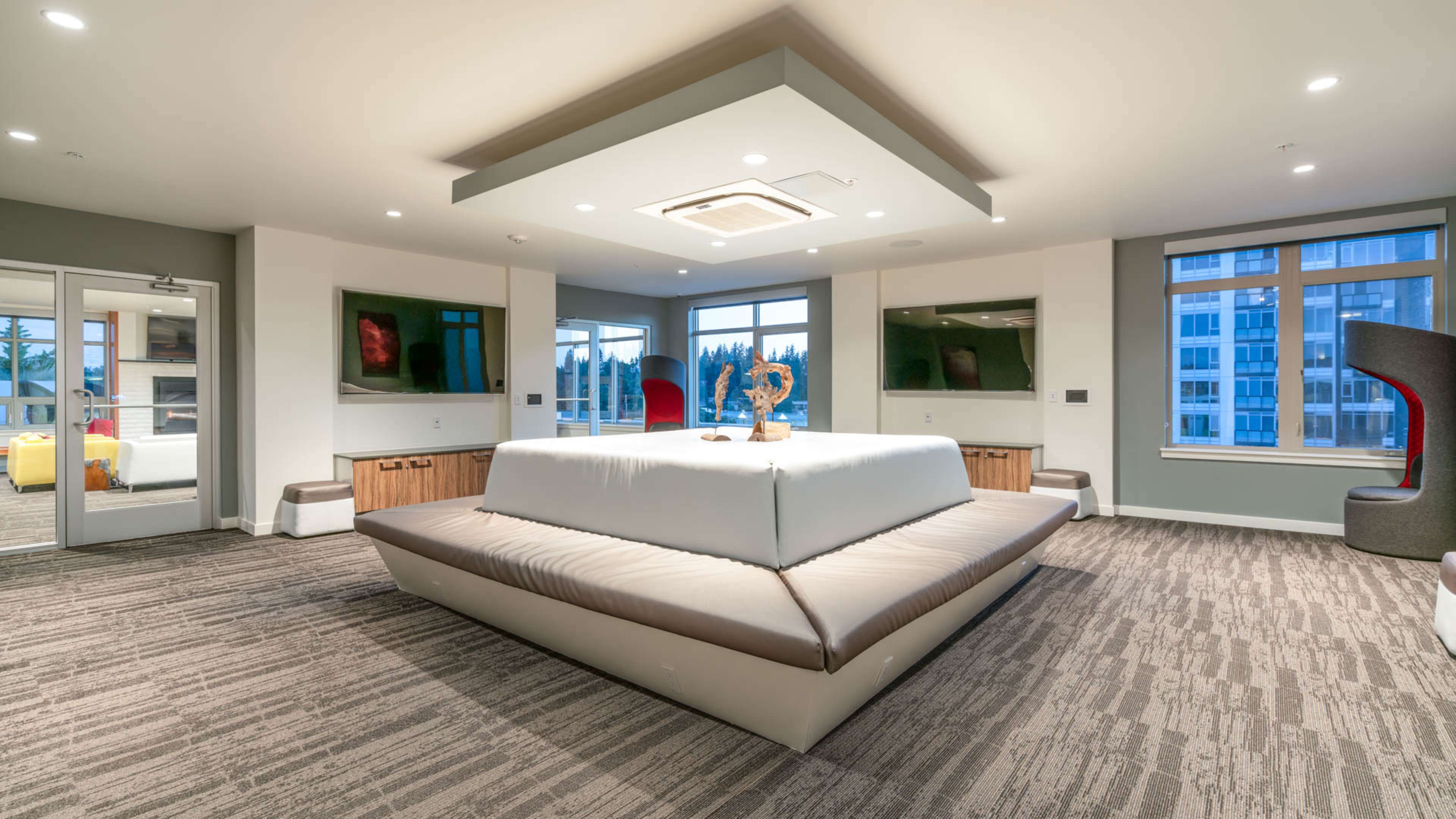 A modern lounge area featuring a large, white cushioned seating arrangement at the center, surrounded by large windows and subtle decor.