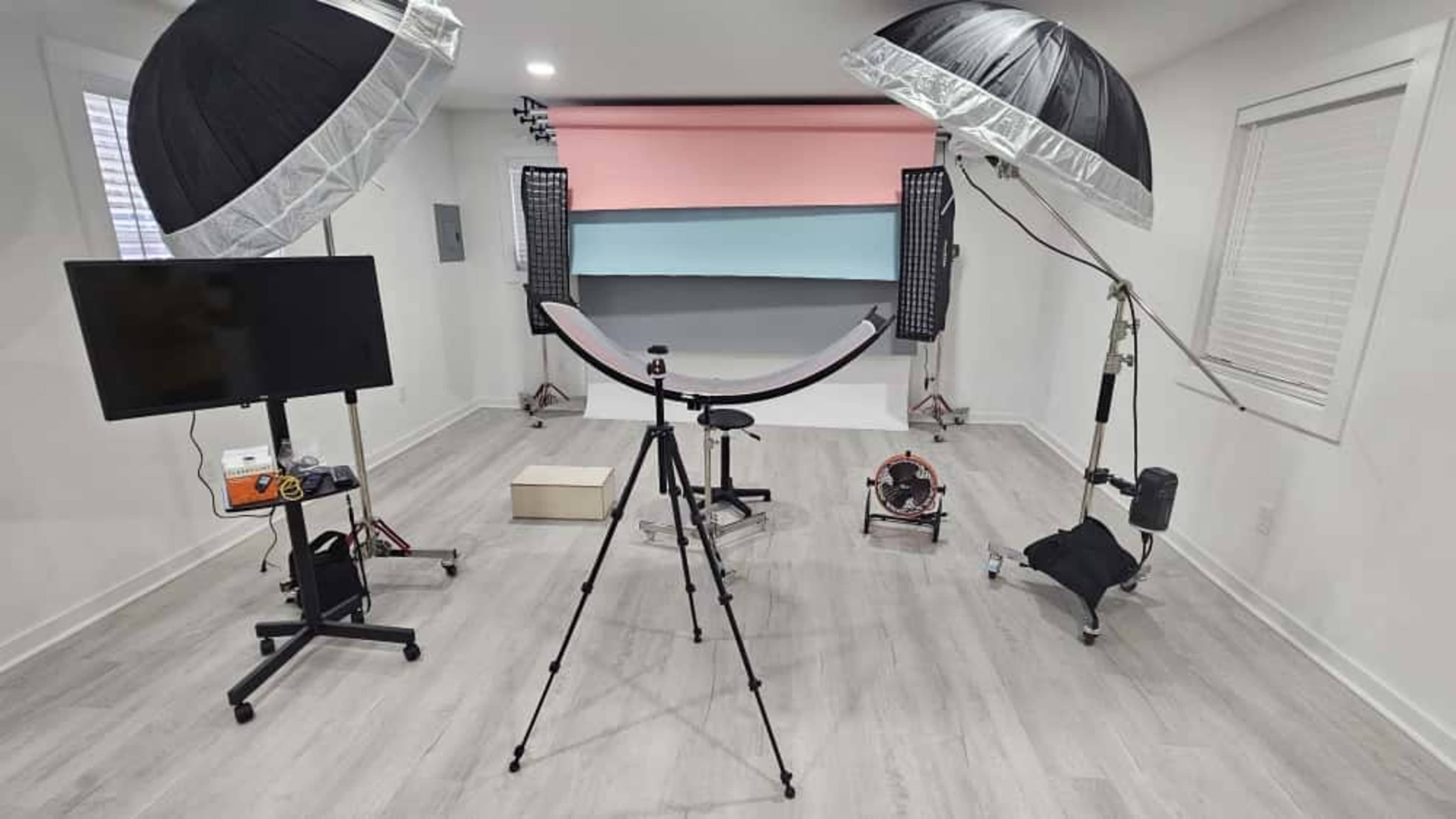 The image shows a photography studio setup with two softboxes, equipment stands, and a background featuring painted colors, along with a tripod and a television monitor.