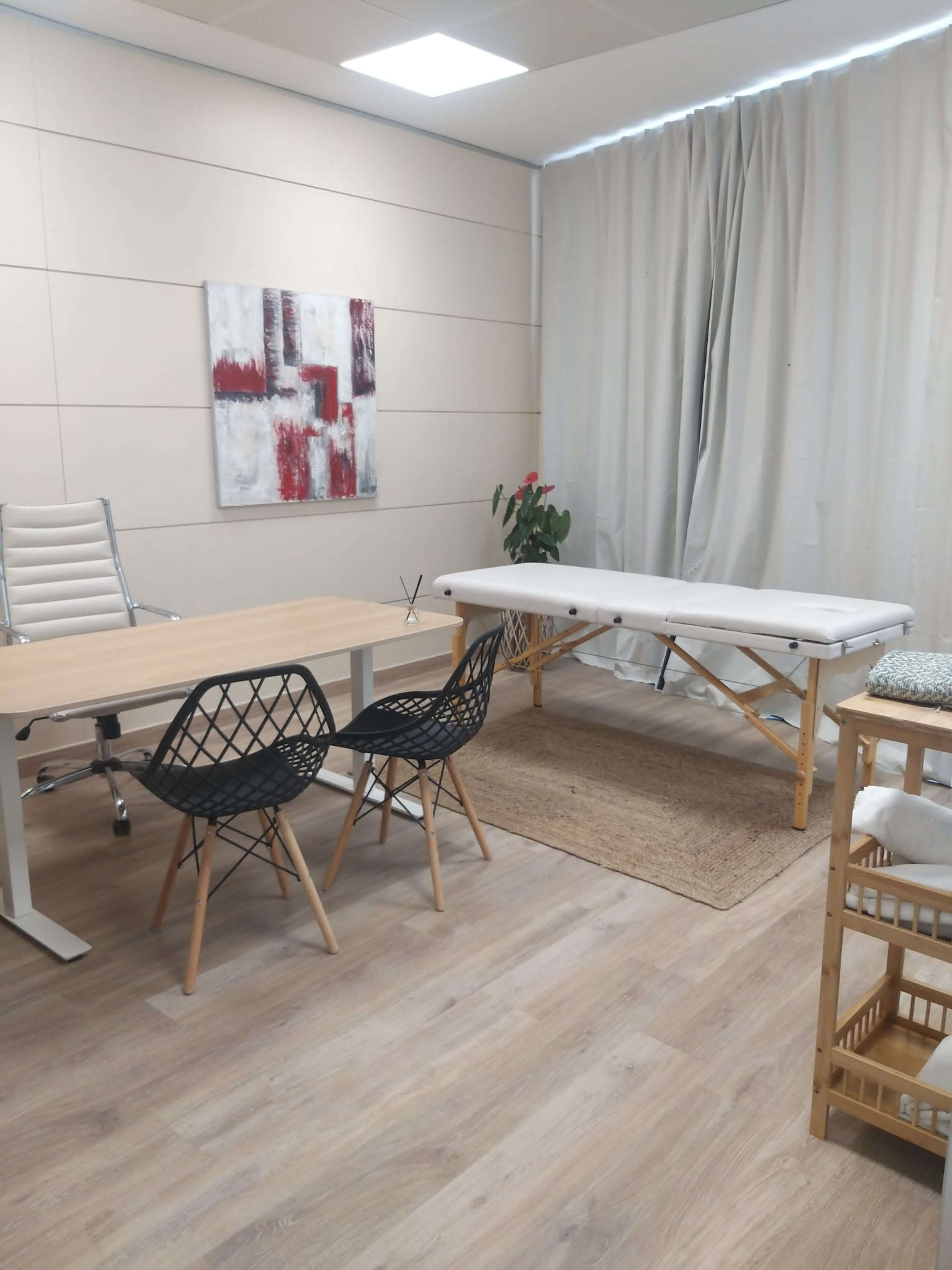 A small, well-organized room features a wooden table with two black chairs, a massage table, and a plant, with light-colored walls and curtains.