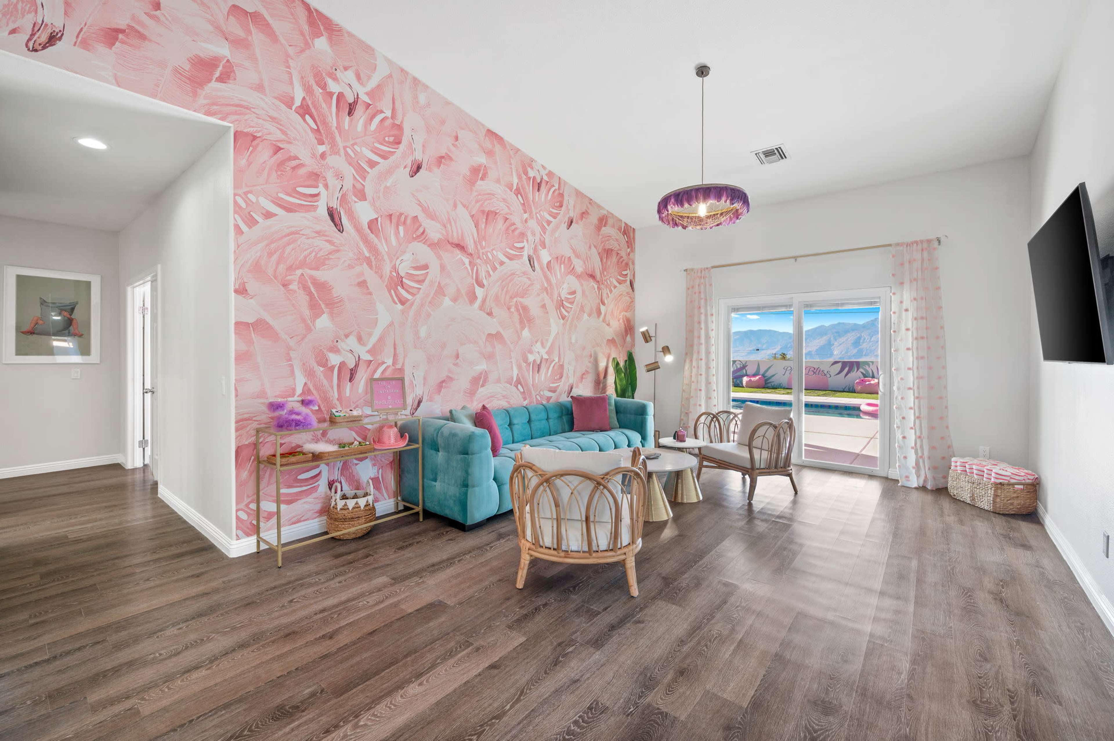 A brightly decorated living room features a pink floral accent wall, turquoise sofa, and wooden furniture, with large windows providing a view of the mountains.