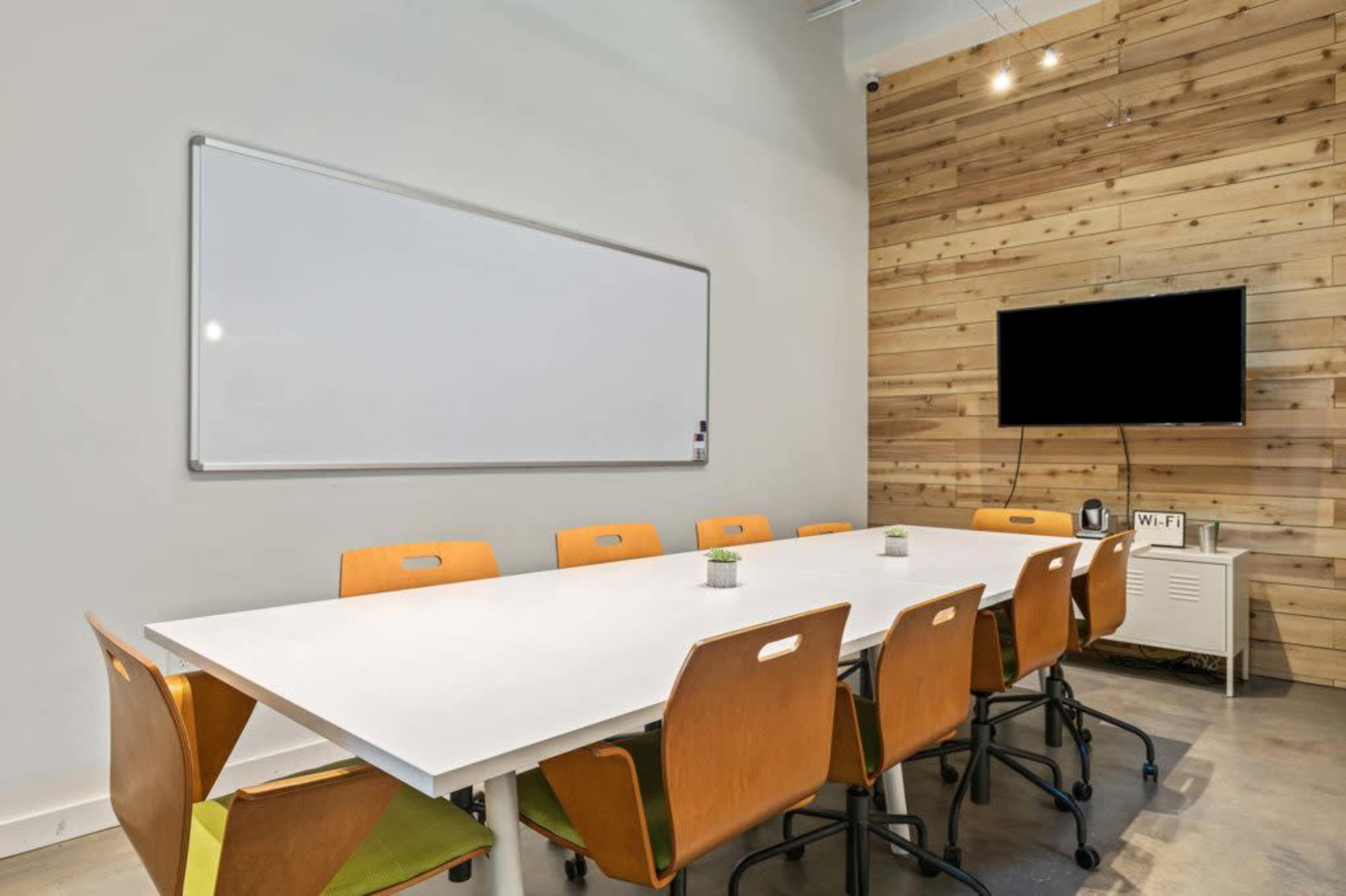 A modern conference room features a long white table with eight wooden chairs, a whiteboard on the wall, a television mounted nearby, and a Wi-Fi signal indicator.