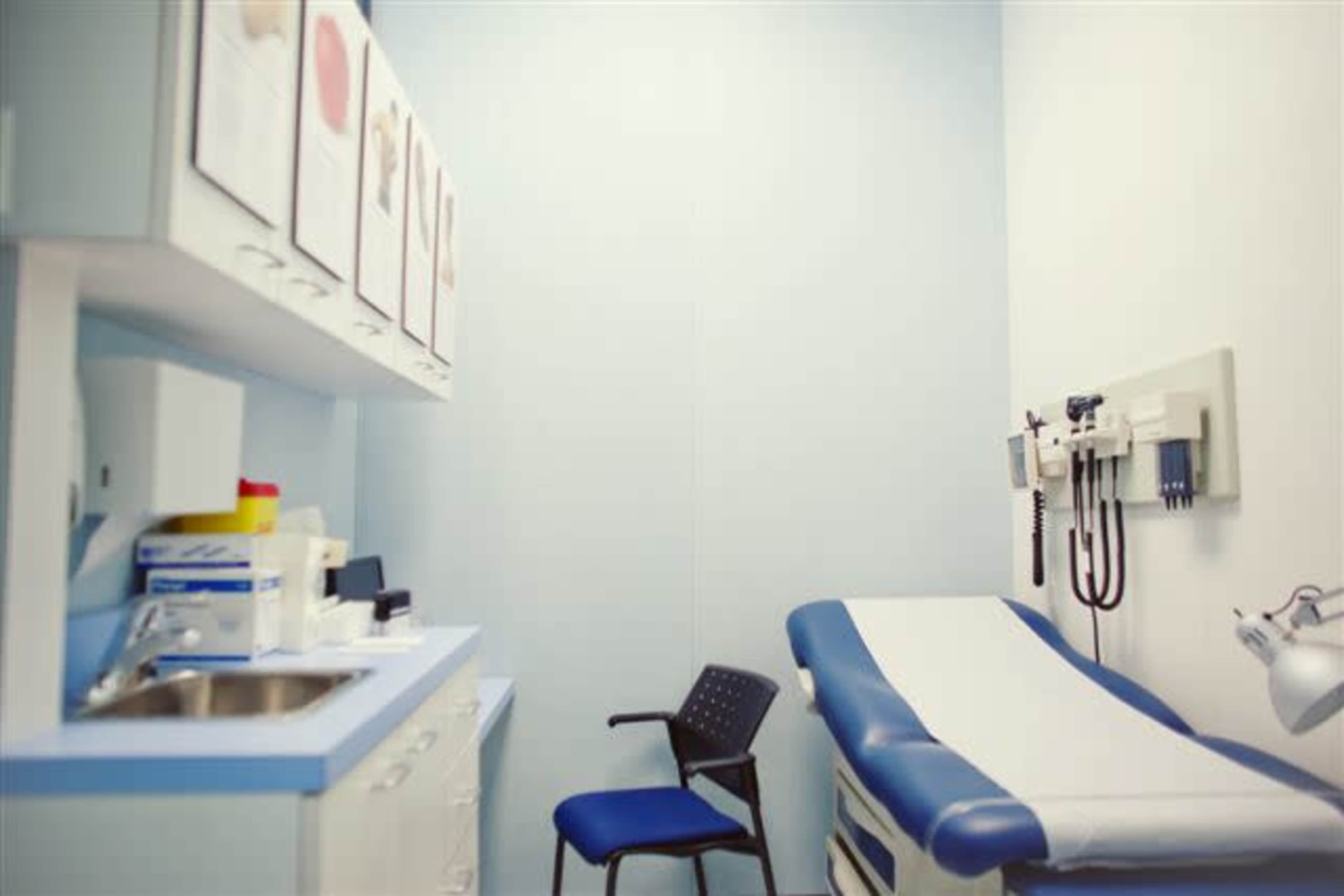 A medical examination room featuring a blue examination table, a chair, and wall-mounted medical equipment.