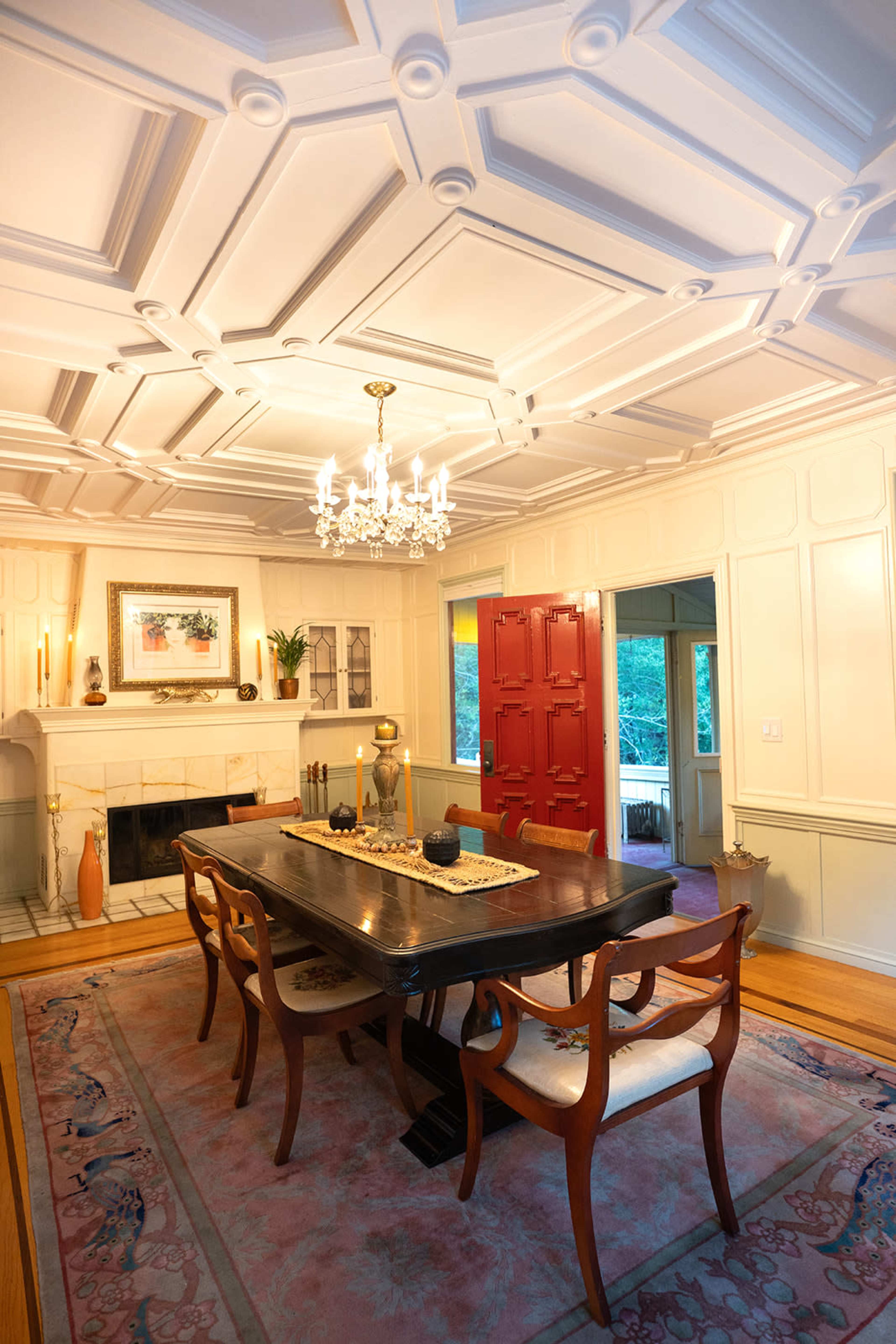 The image shows a dining room with a coffered ceiling, a long dark wooden table surrounded by four chairs, and a red front door in the background.