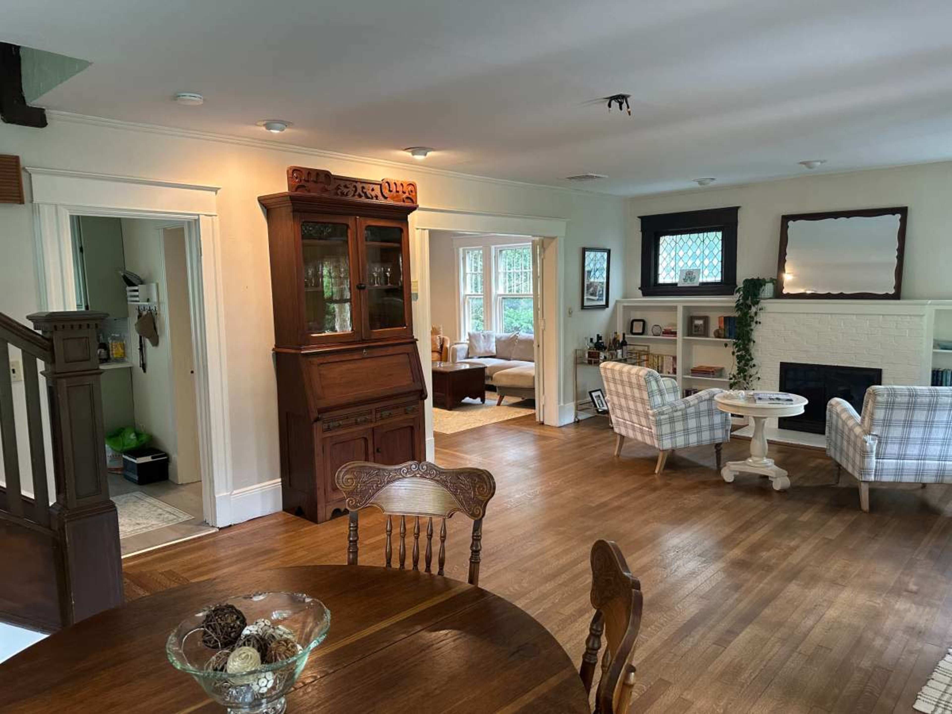 The image shows a spacious, well-lit interior of a home featuring a dining area with a wooden table, a decorative china cabinet, and a living area with two armchairs and a fireplace.