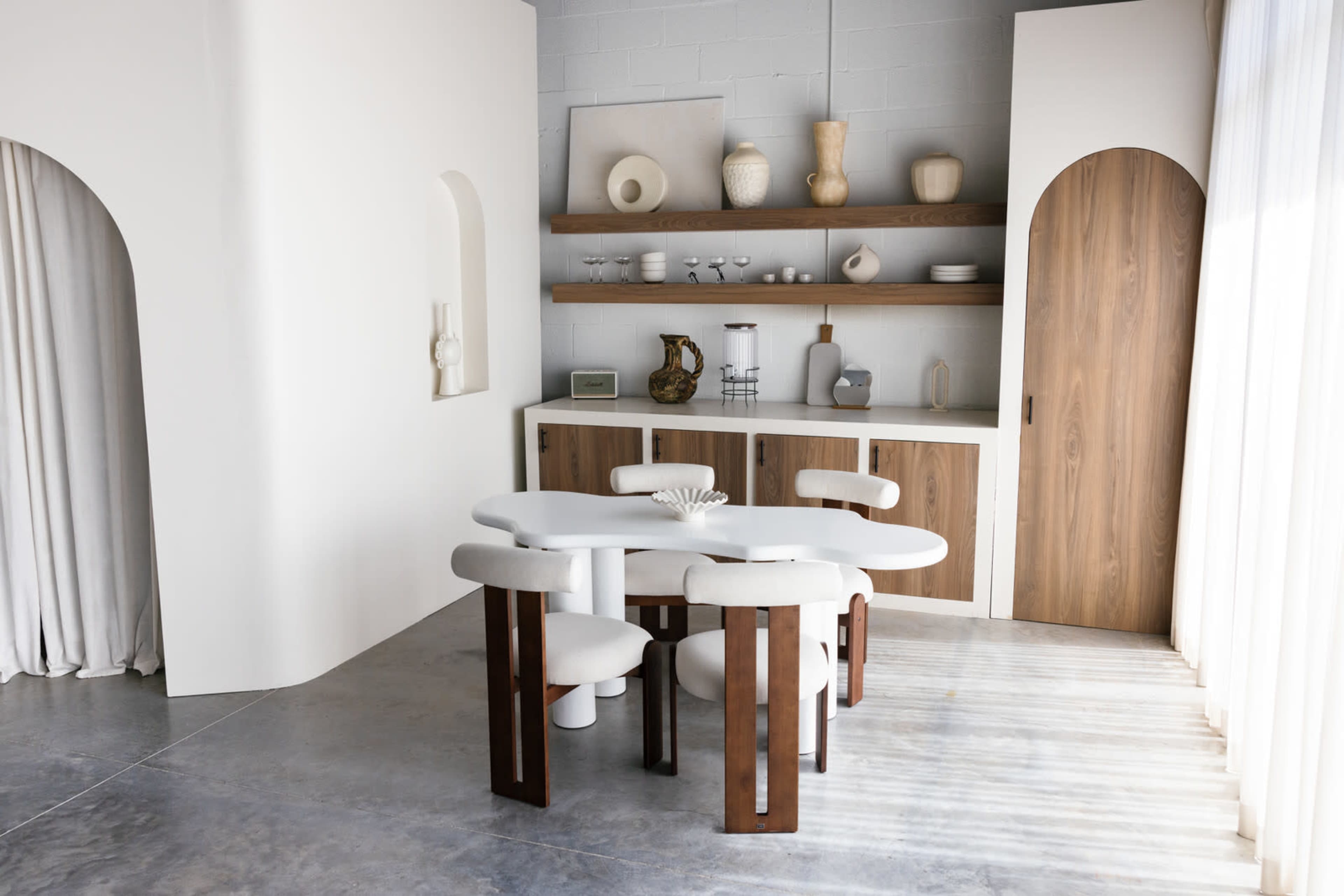 A modern dining area featuring a curved table with four upholstered chairs, a wooden shelf displaying decorative items, and a cabinet with a wooden door.