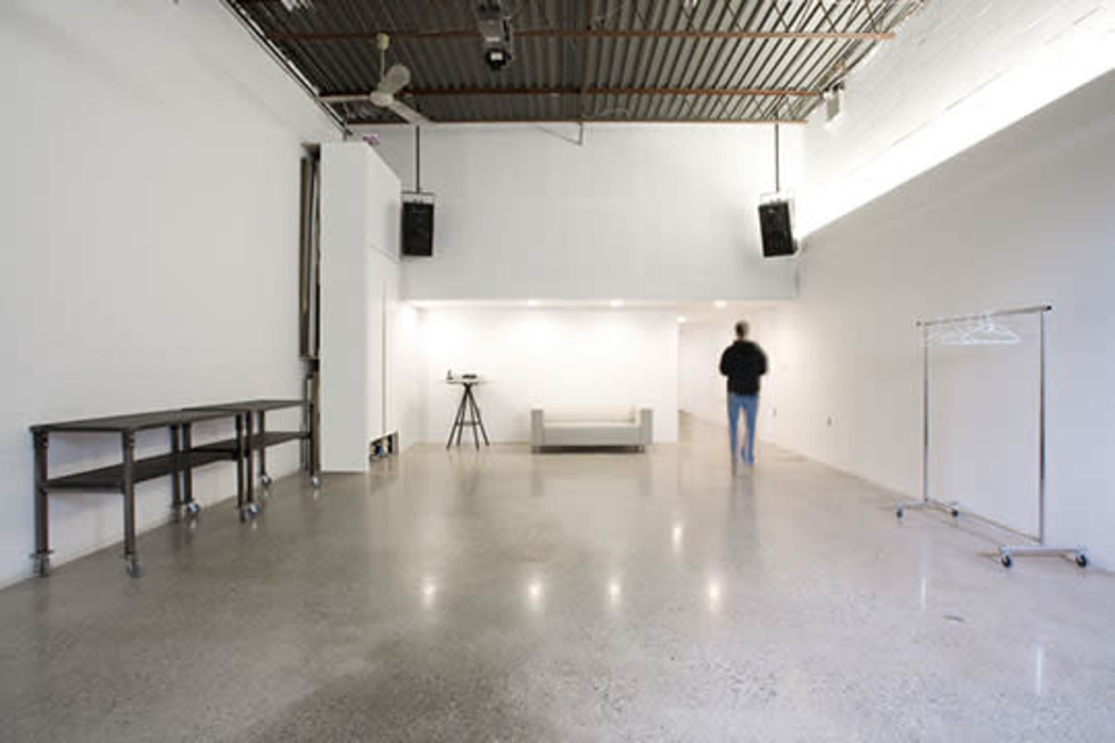 The image shows a spacious, minimalist room with polished concrete flooring, a sofa in the center, a standing clothing rack, and a person walking towards the back of the room.