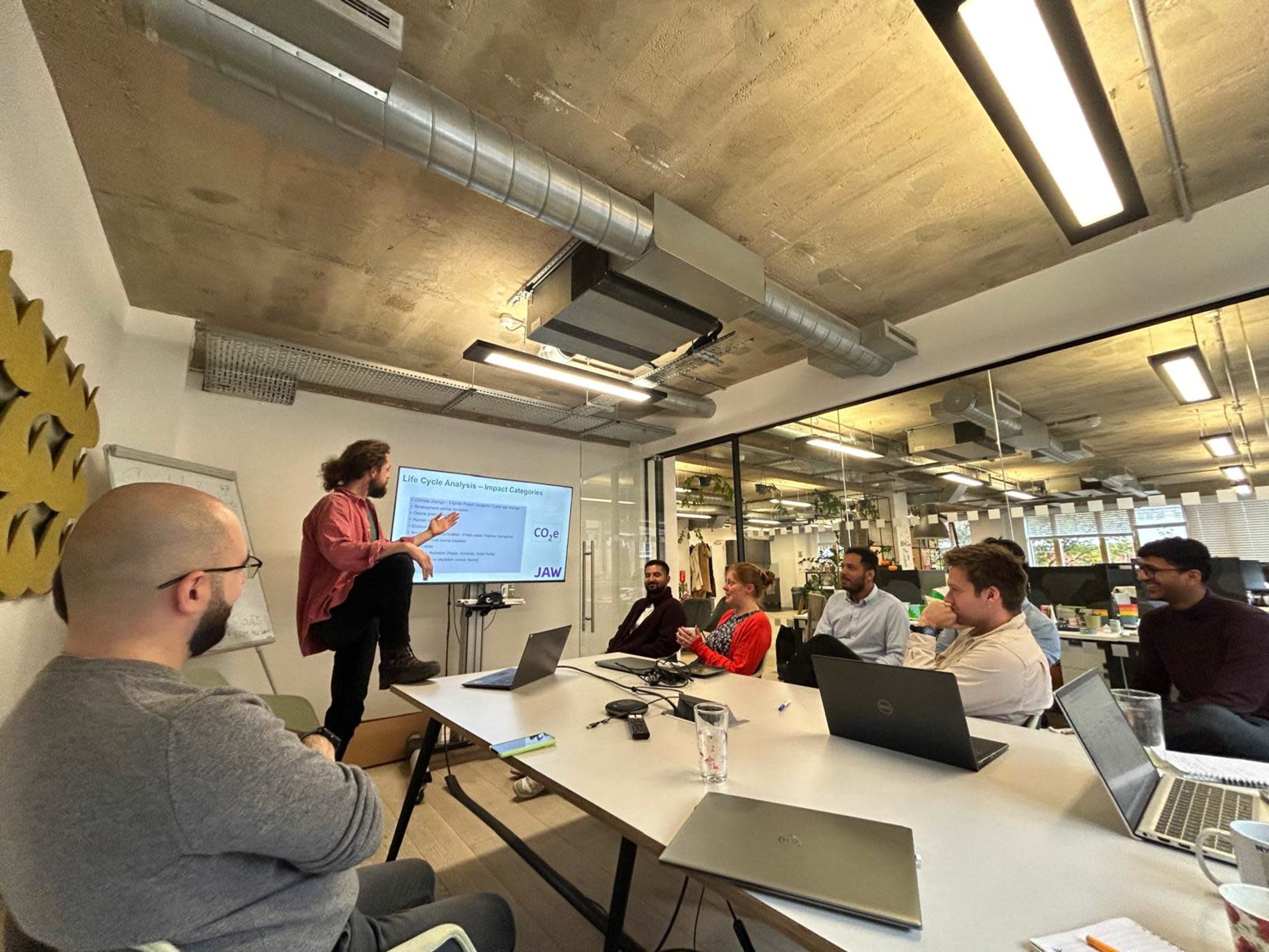 A man is presenting a presentation in a modern office meeting room while seated attendees look on.