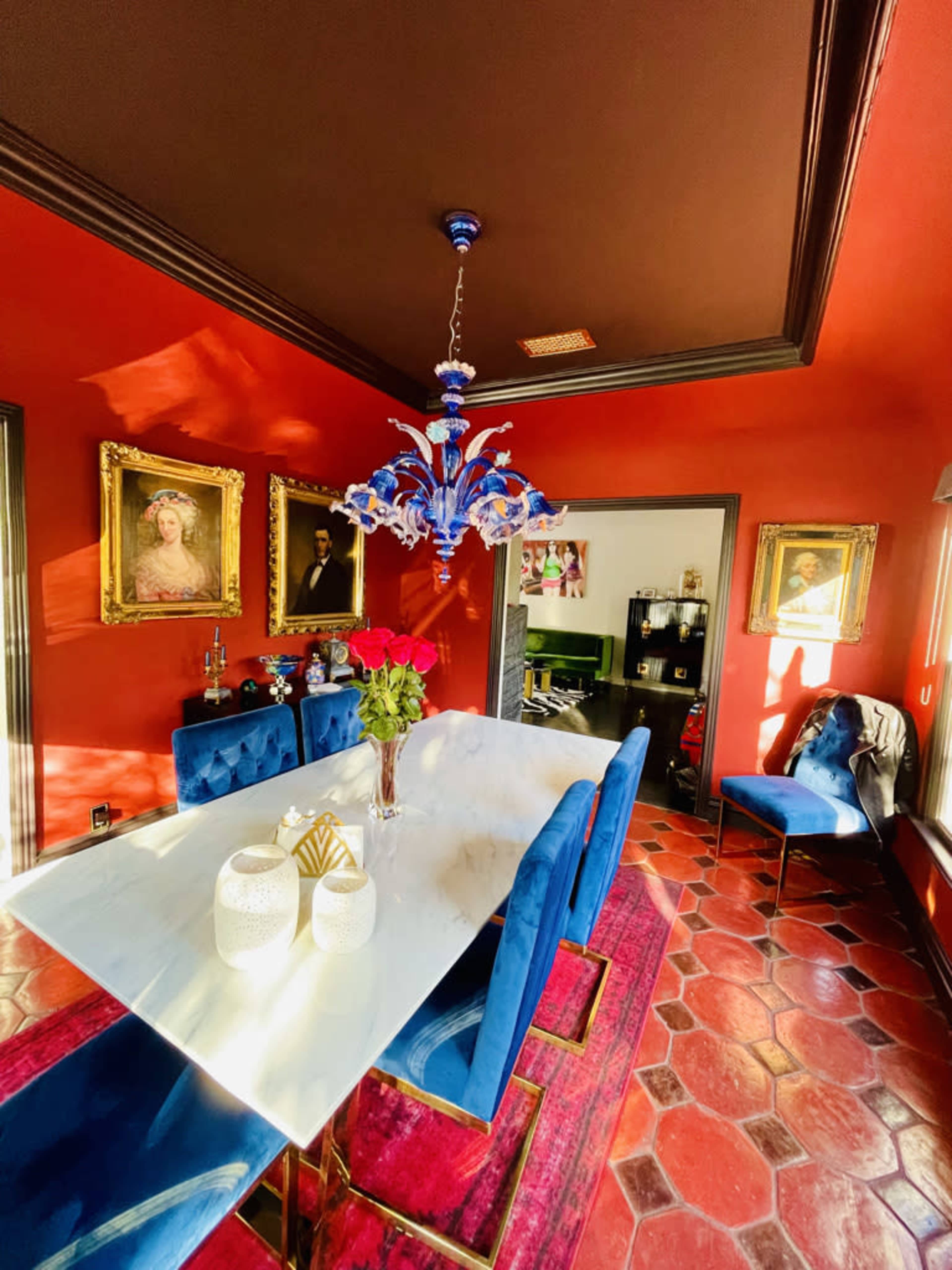 A dining room features a marble table surrounded by blue upholstered chairs, with a colorful chandelier and portraits on the red walls.