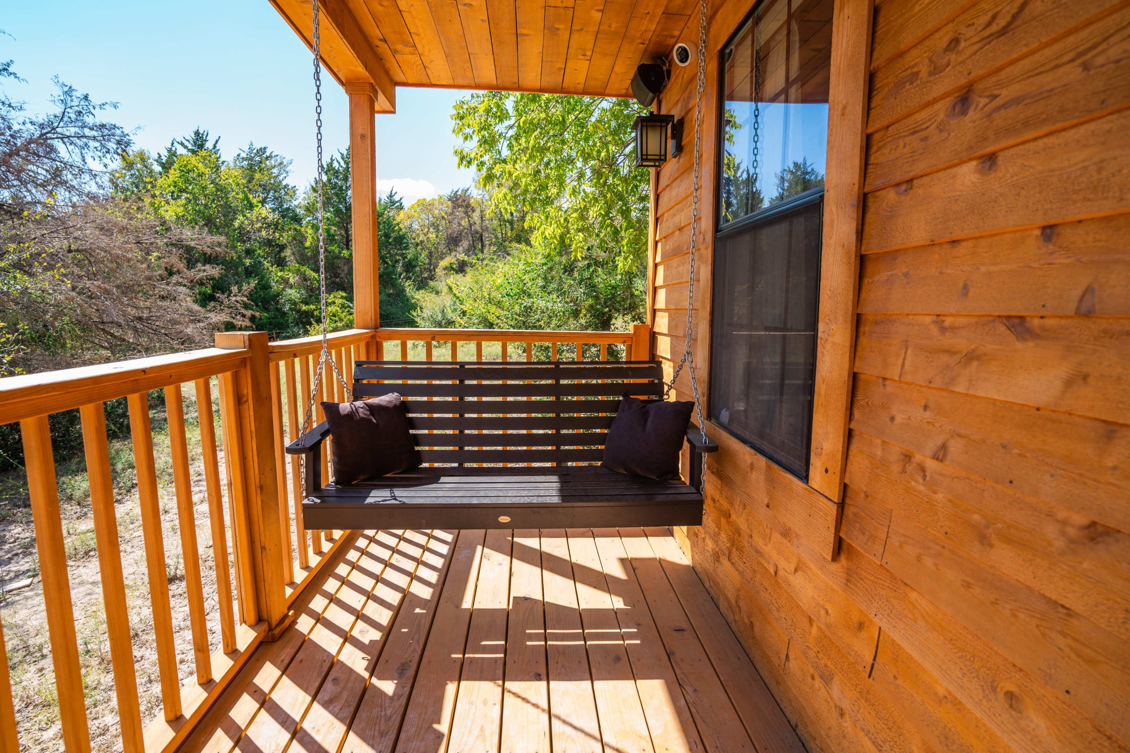 A wooden porch with a swing and two pillows, surrounded by greenery.