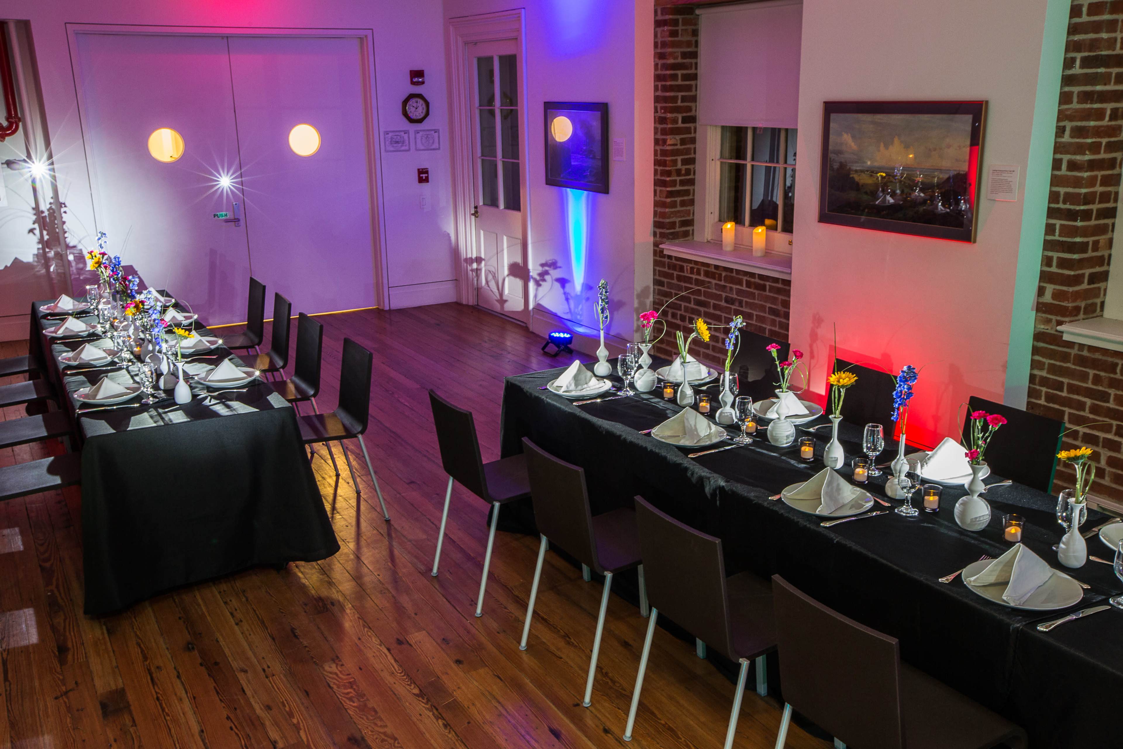 A setup for a dining event featuring two long tables adorned with decorative tableware and flowers, under soft colored lighting in a brick-walled room.
