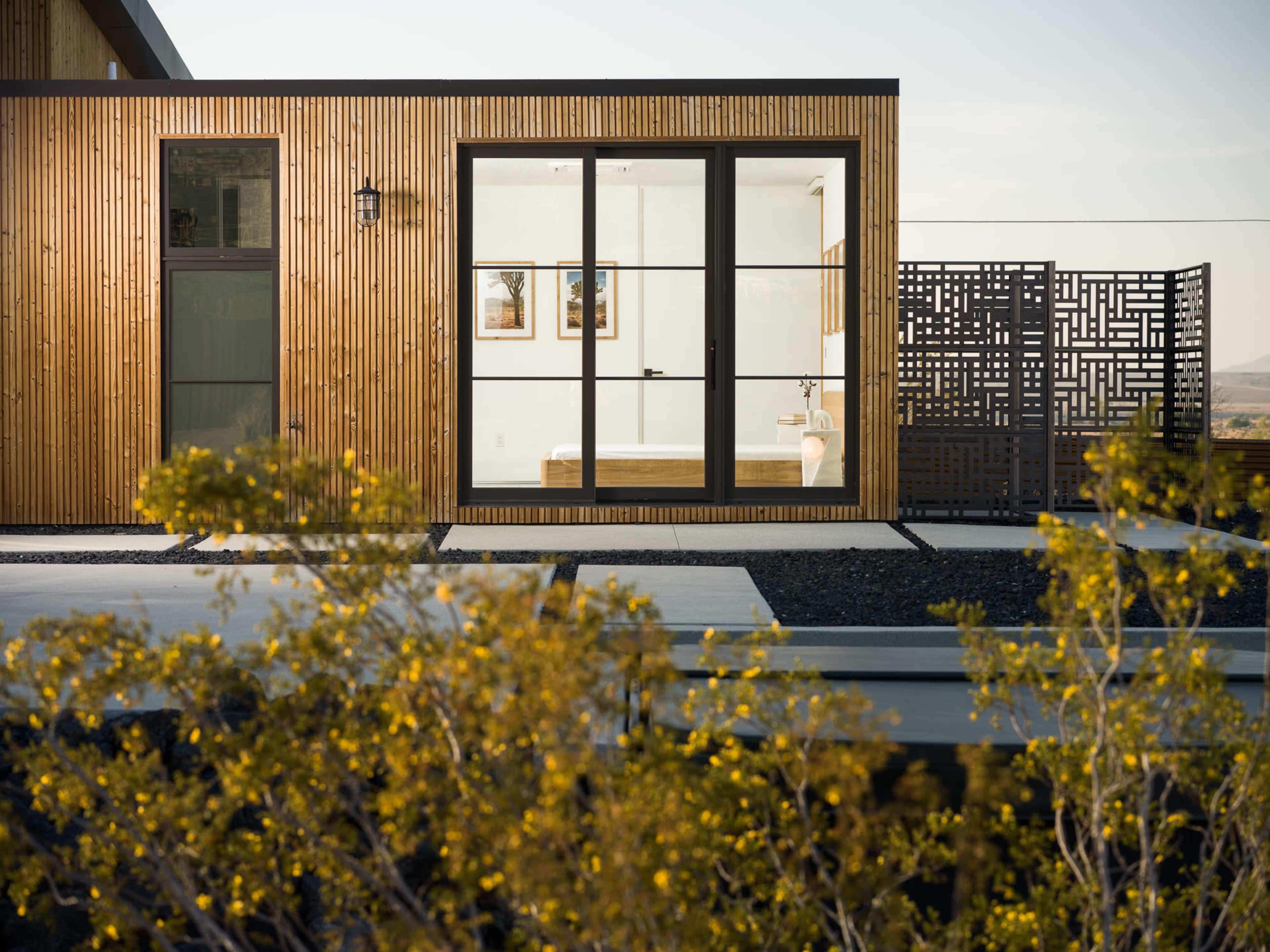 A modern wooden building features large windows displaying a bathroom, with a decorative screen and yellow foliage in the foreground.