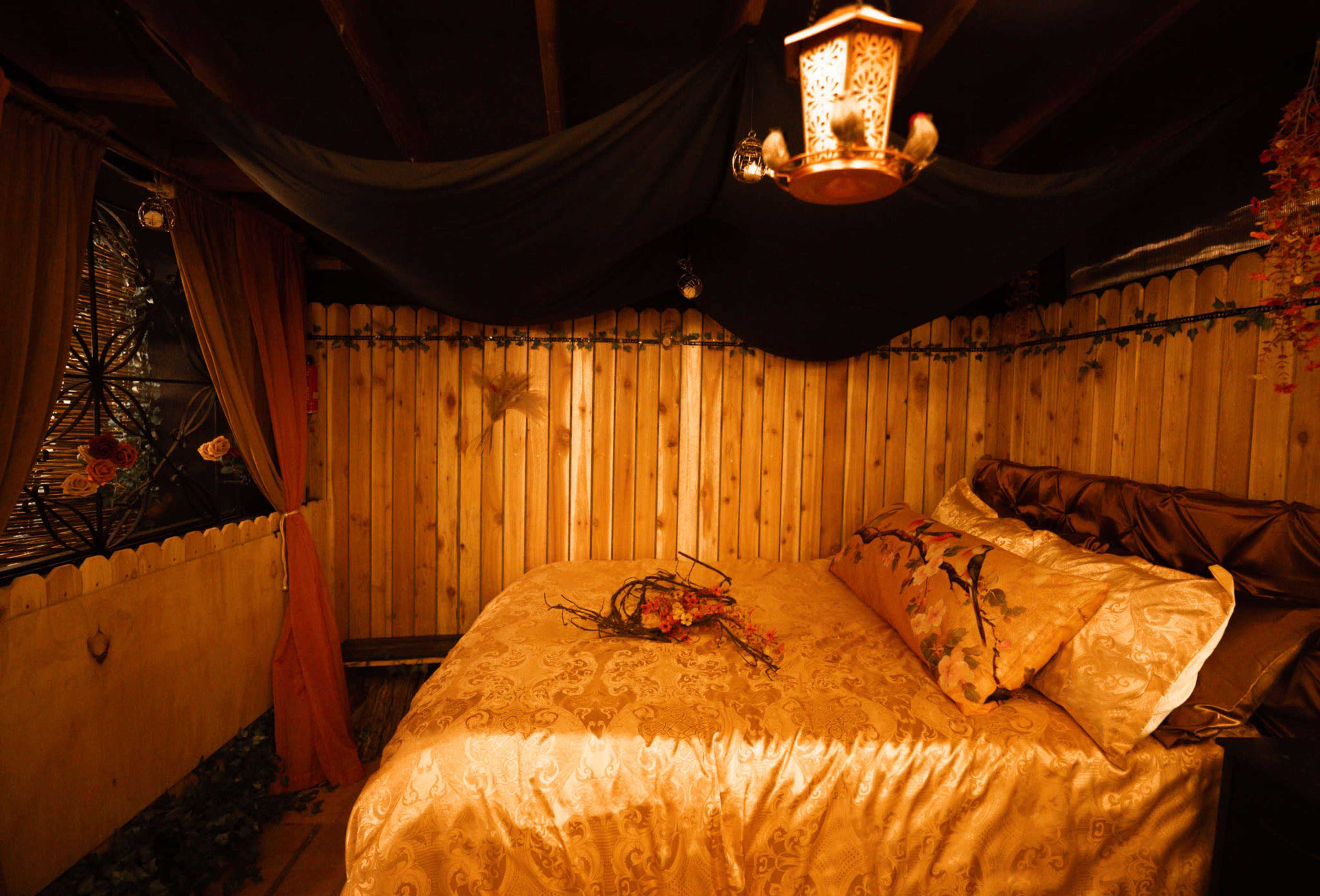 The image depicts a cozy wooden room with a bed adorned in golden bedding and decorative pillows, surrounded by rustic wooden walls and soft ambient lighting.
