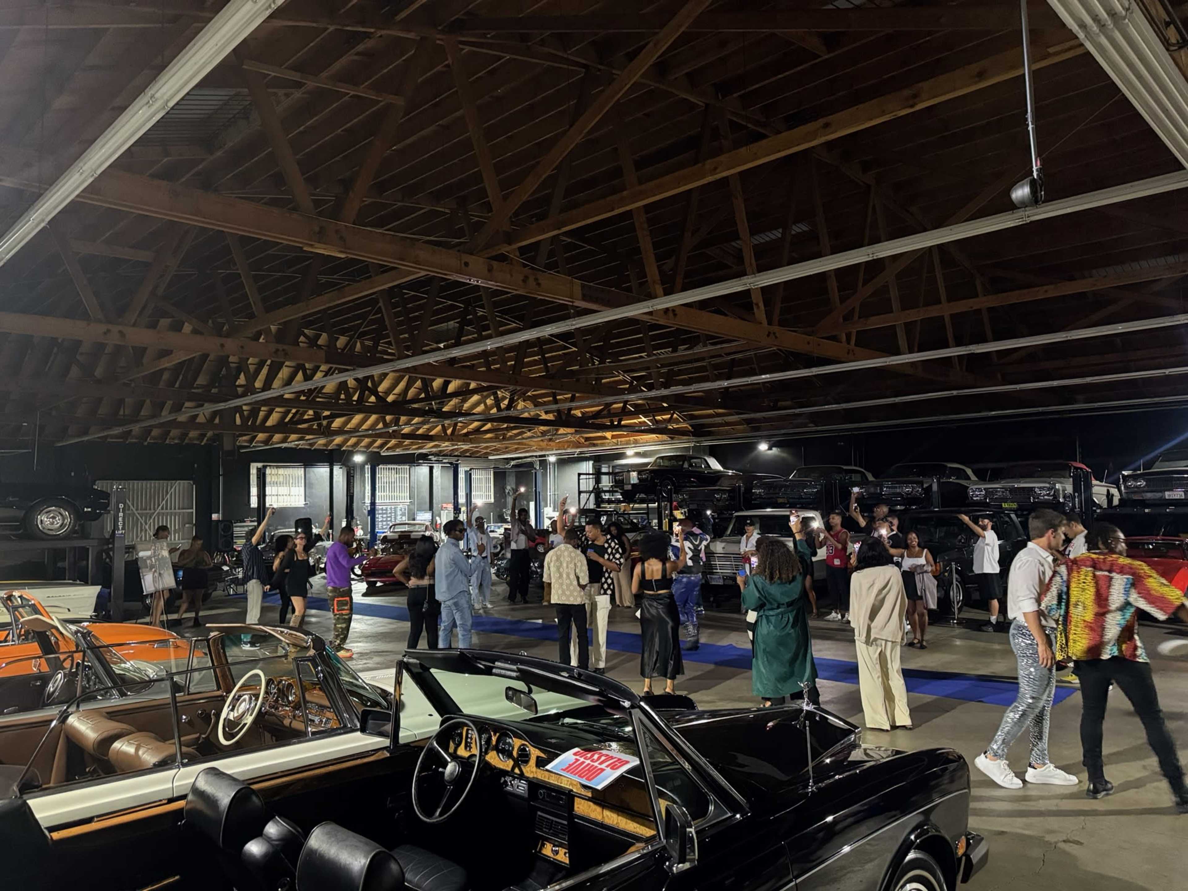 A crowd of people mingles in a spacious venue filled with vintage cars under a wooden ceiling.