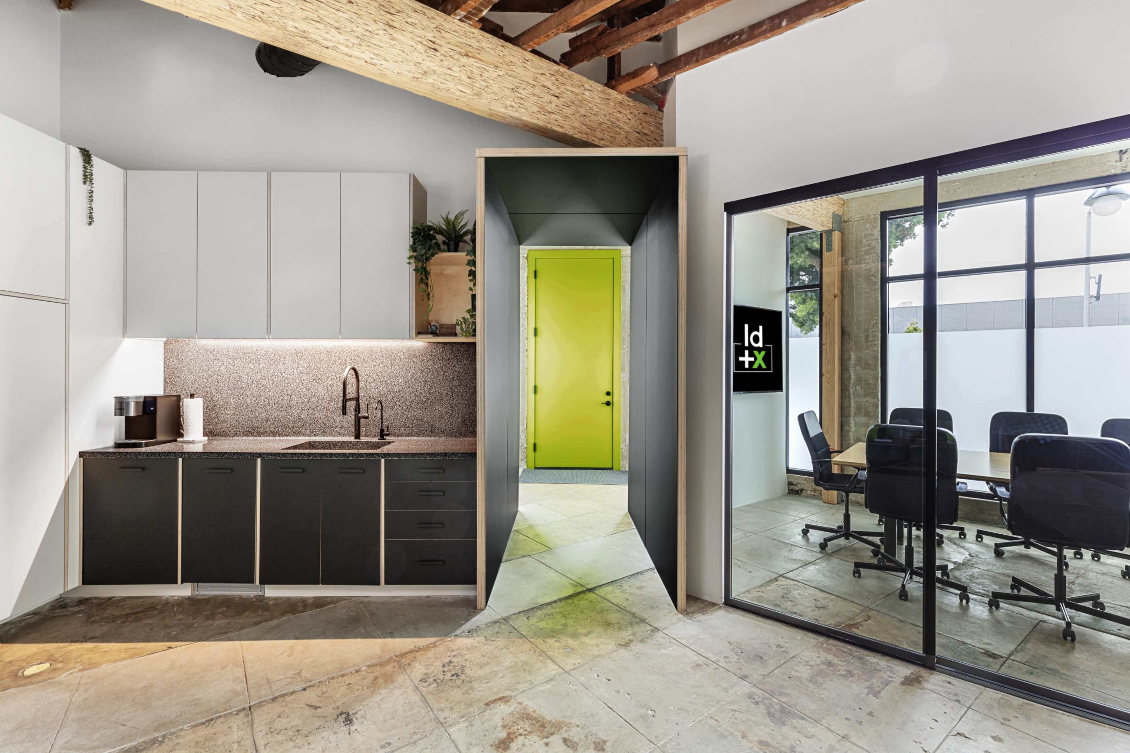 The image shows a modern office space featuring a kitchenette on the left, an entry to a brightly colored room in the center, and a glass-walled conference area on the right.