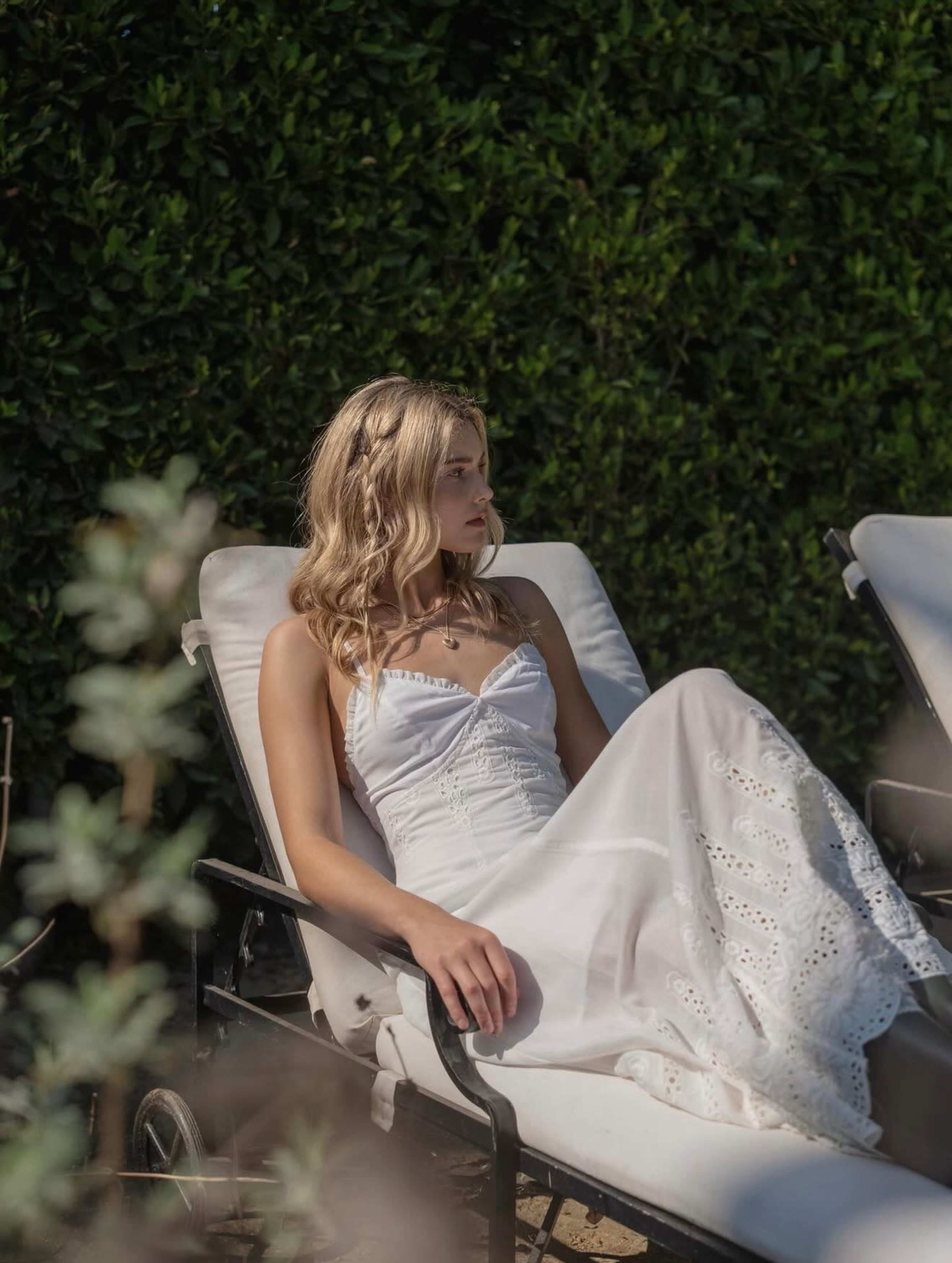 A young woman in a white dress reclines on a lounge chair in a garden setting.