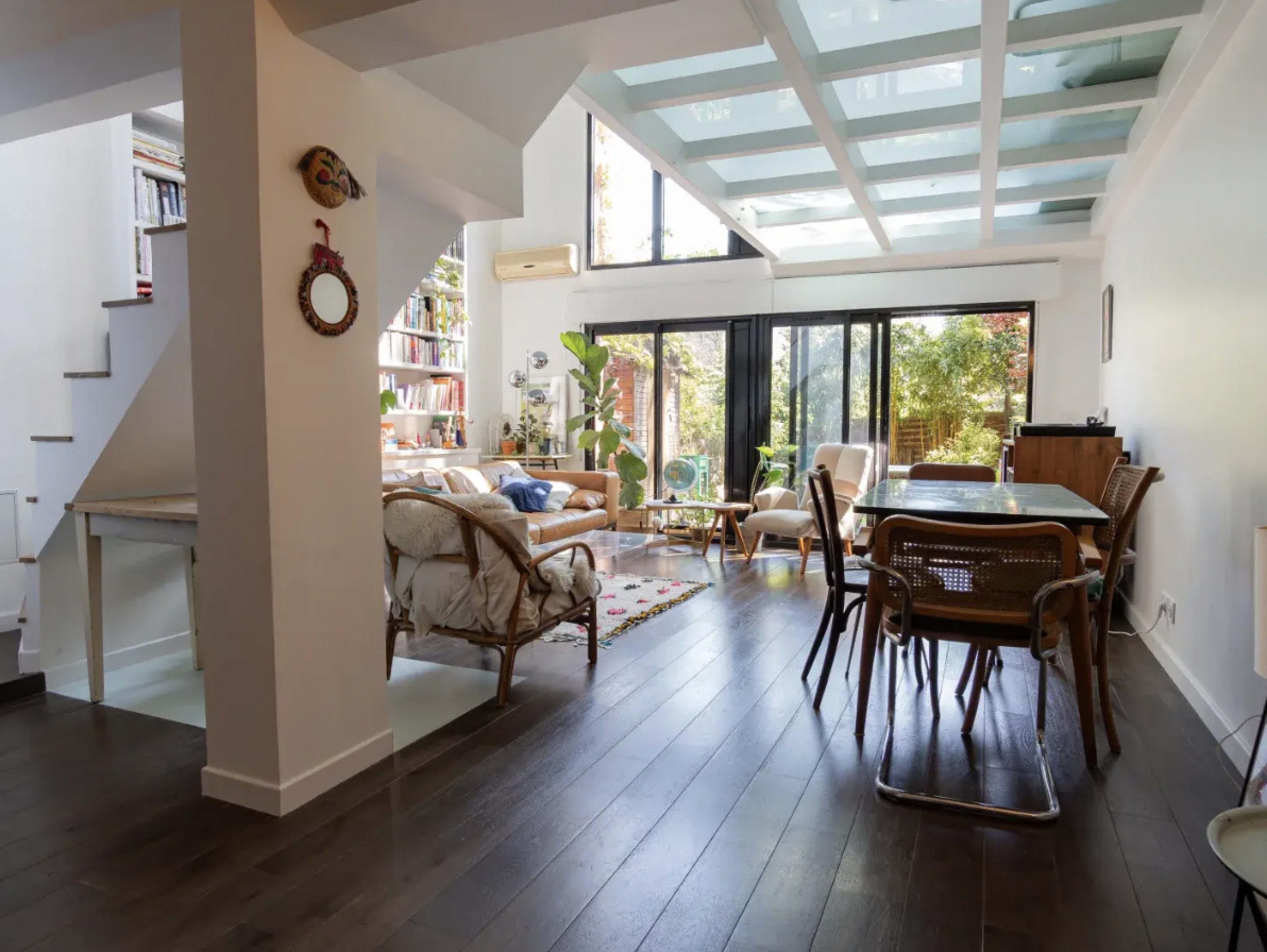The image shows a modern, open-concept living and dining area with large windows, offering views of a garden outside.