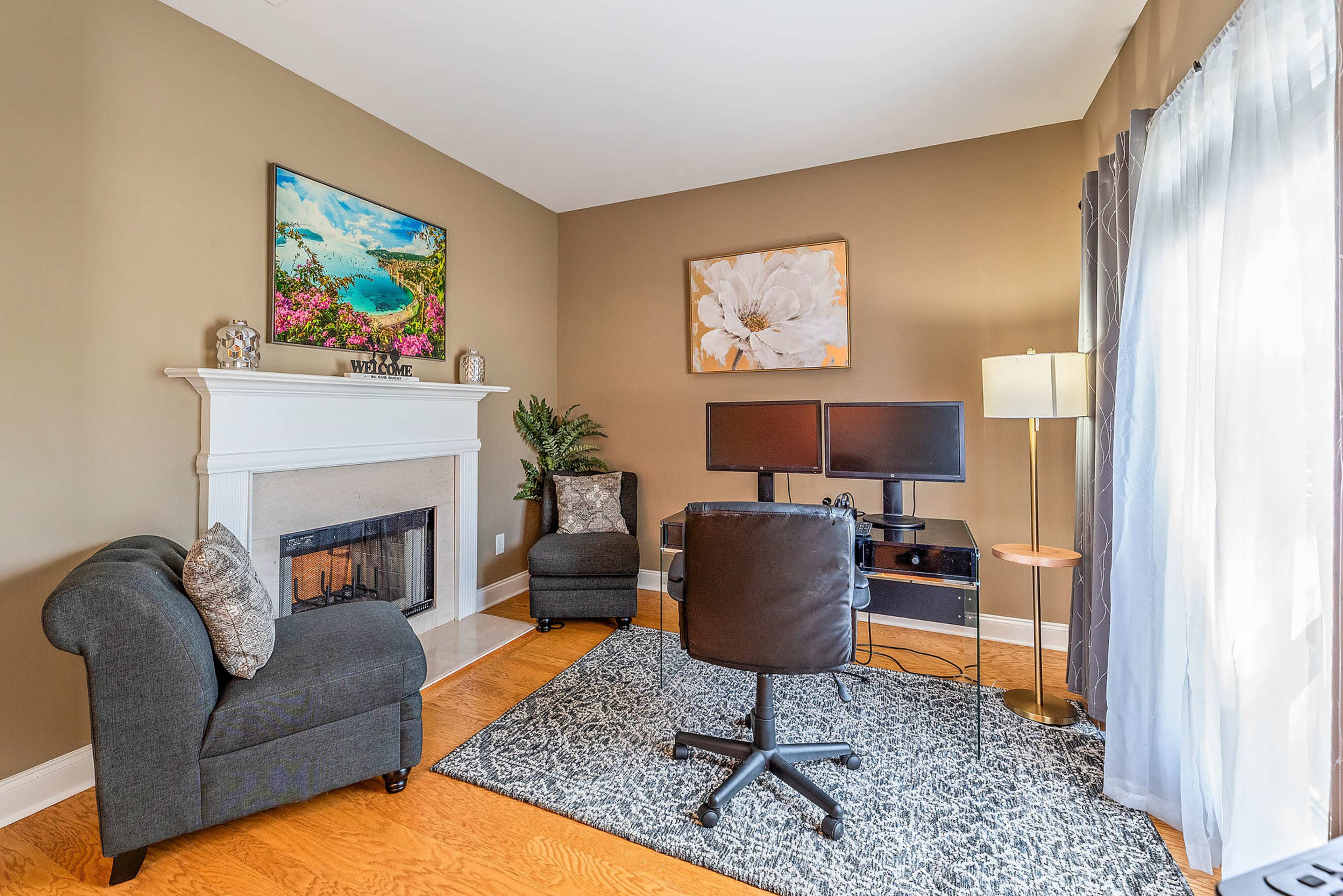 A cozy home office featuring two computer monitors, a comfortable chair, and a decorative fireplace, all set against a neutral wall.