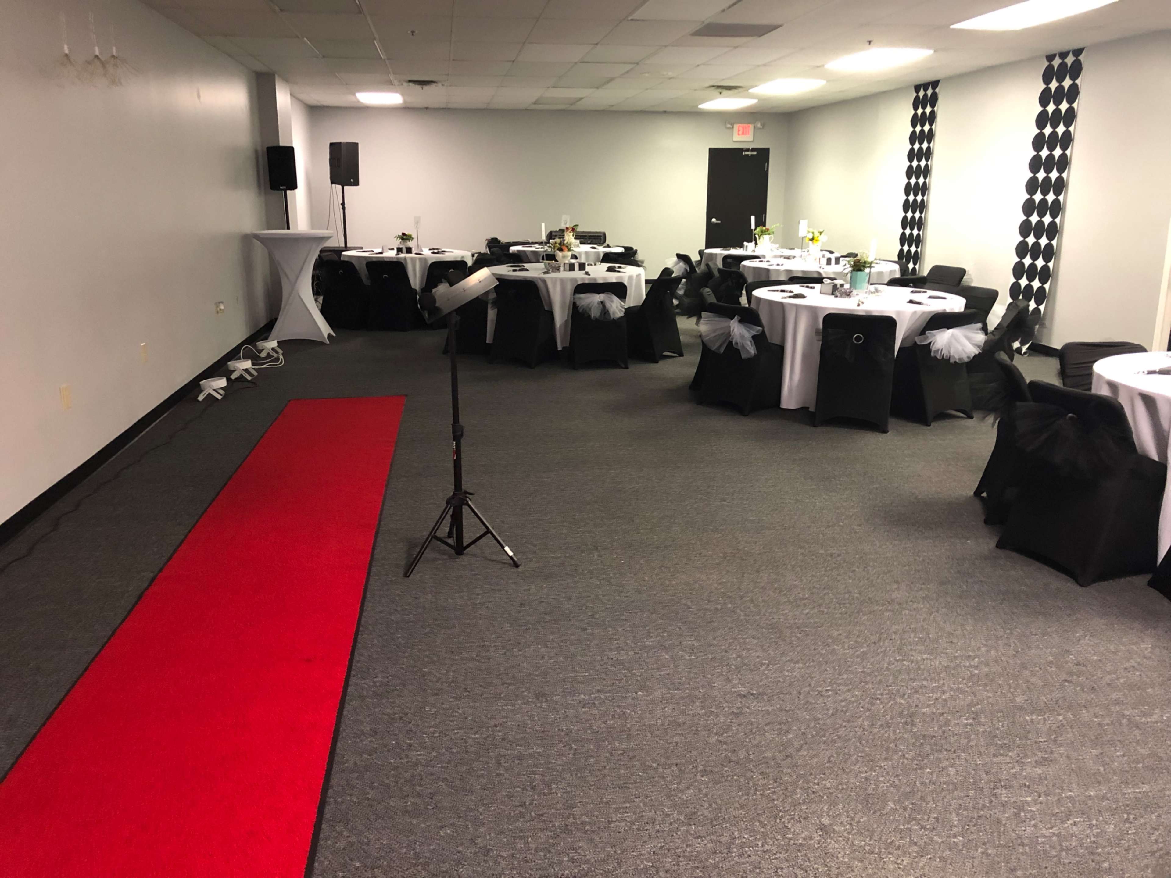 The image shows a decorated event space with several tables set with white tablecloths and black chairs, alongside a red carpet leading to a microphone stand.