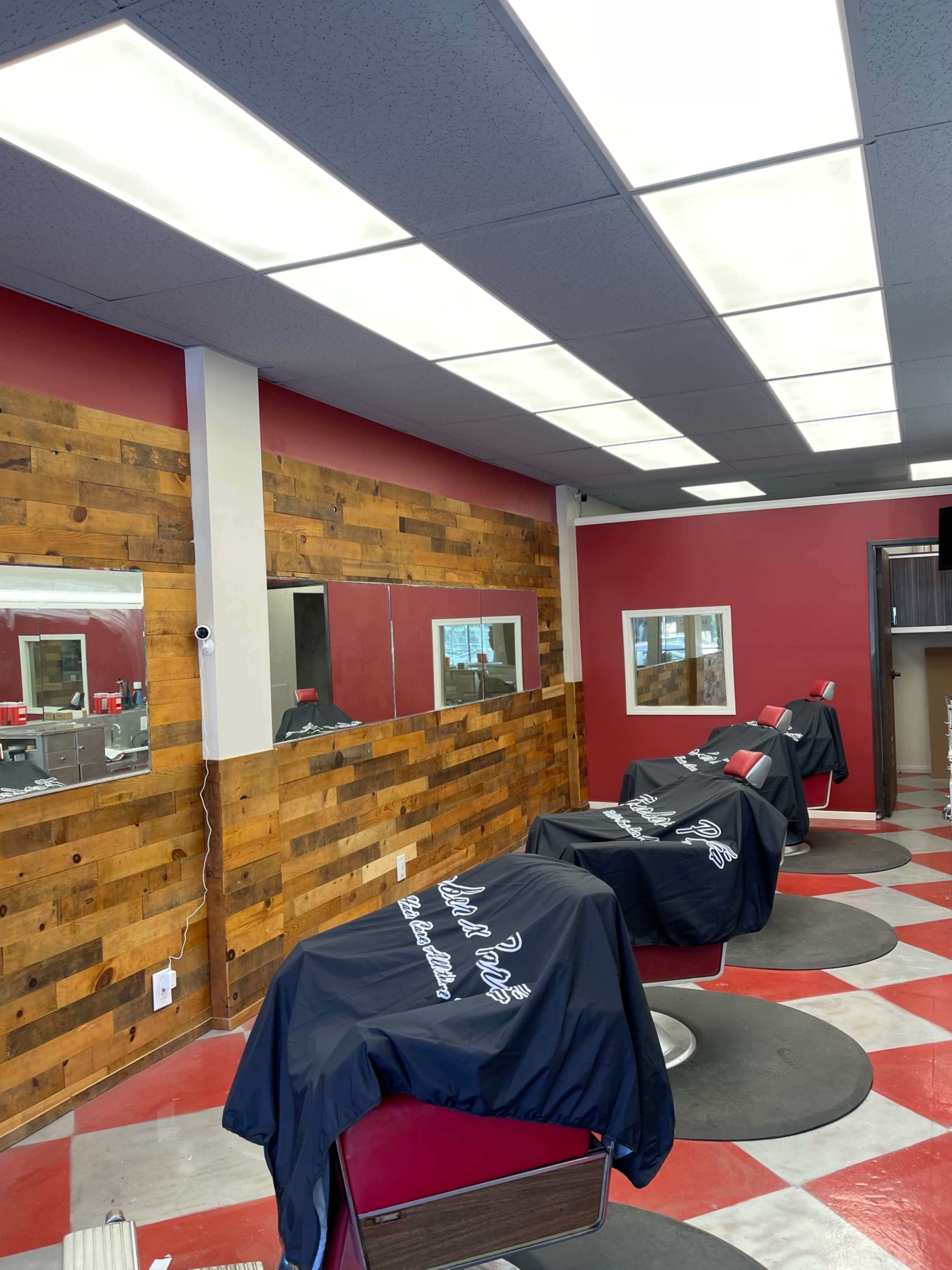 The image shows the interior of a barbershop featuring wooden accent walls, red and white checkered flooring, and several barber chairs covered with black capes.