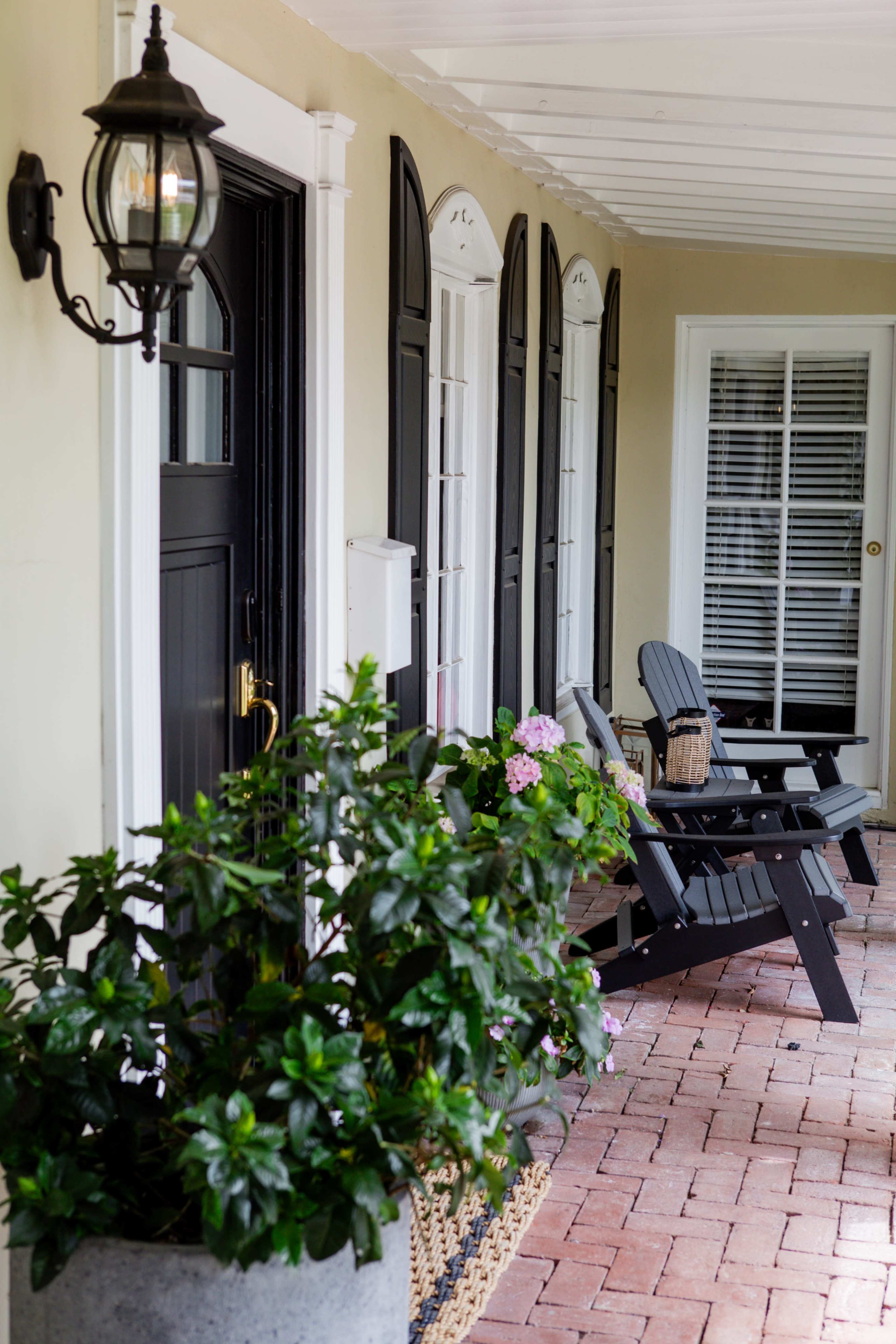 The image shows a porch with two black wooden chairs, flowering plants, and multiple doors, under a well-lit lantern.