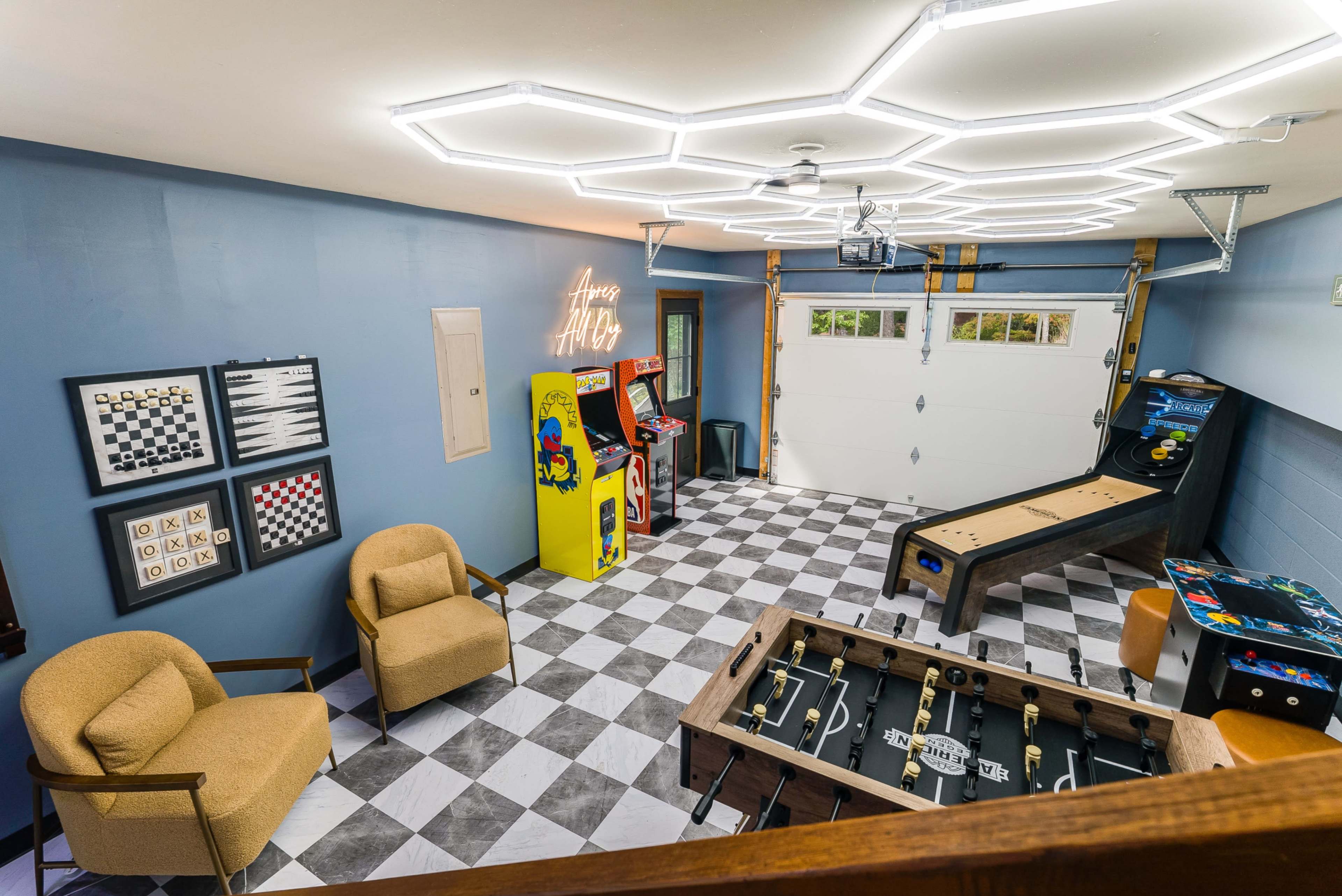The image shows a spacious game room with a patterned floor, featuring arcade machines, a foosball table, a skee ball game, and cozy chairs under modern lighting.