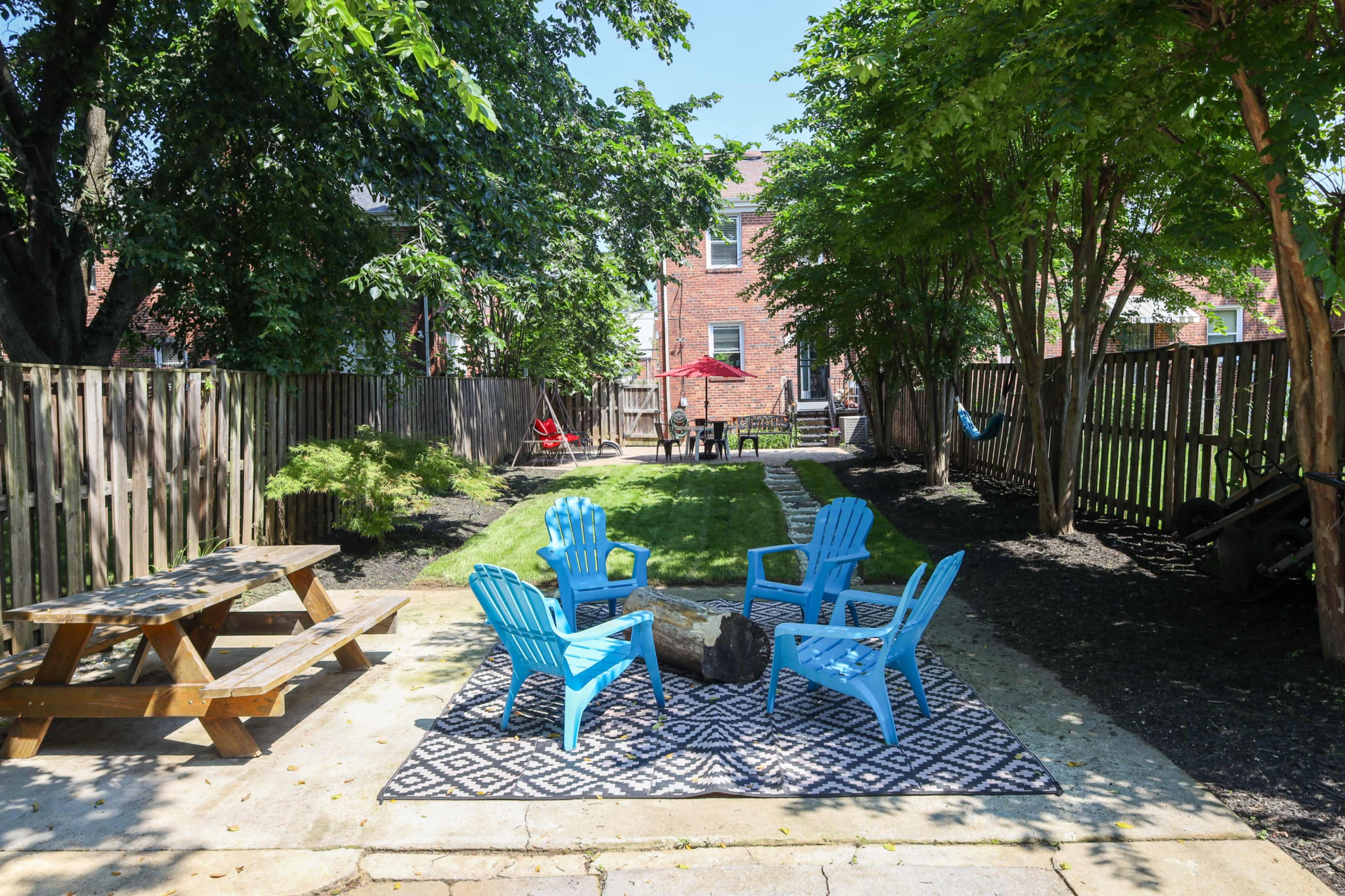 A backyard features a picnic table, four blue chairs arranged around a fire pit, and a grassy area bordered by wooden fences and trees.