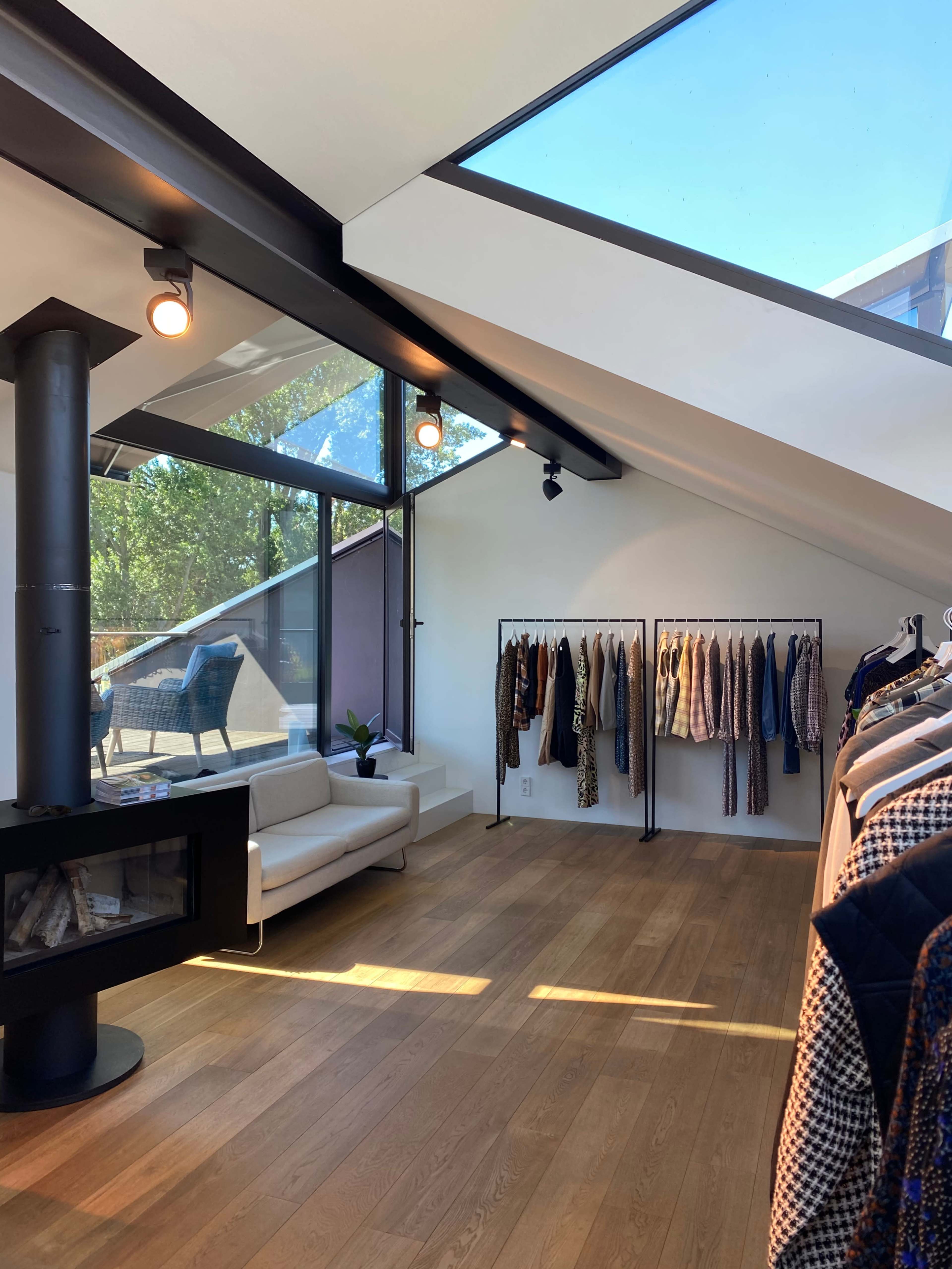 A bright, modern clothing store features a sofa and racks displaying various garments under a sloped ceiling with large windows.