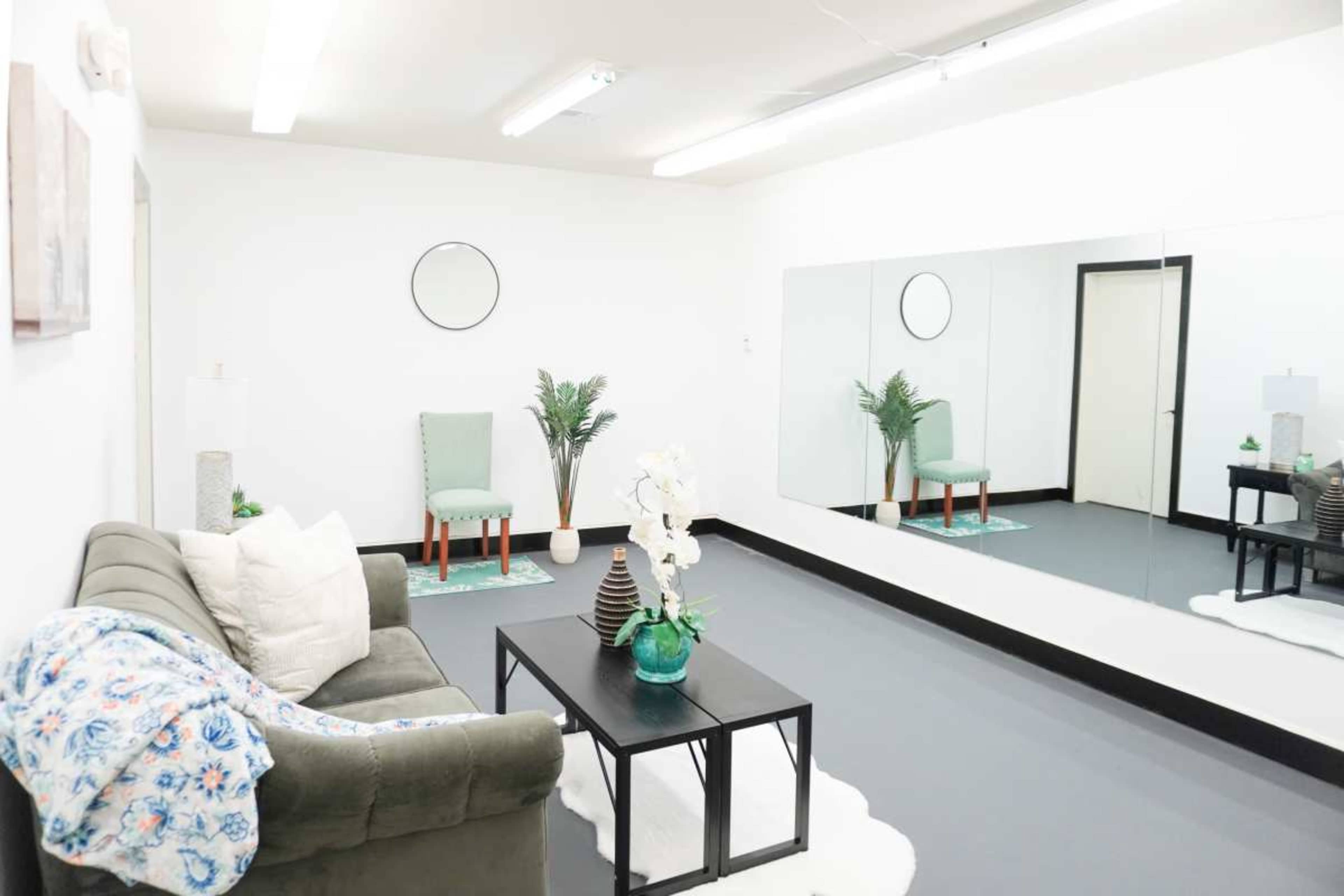 The image shows a minimalist interior space with a green couch, a black coffee table, a round wall mirror, and two chairs against a mirror-clad wall.